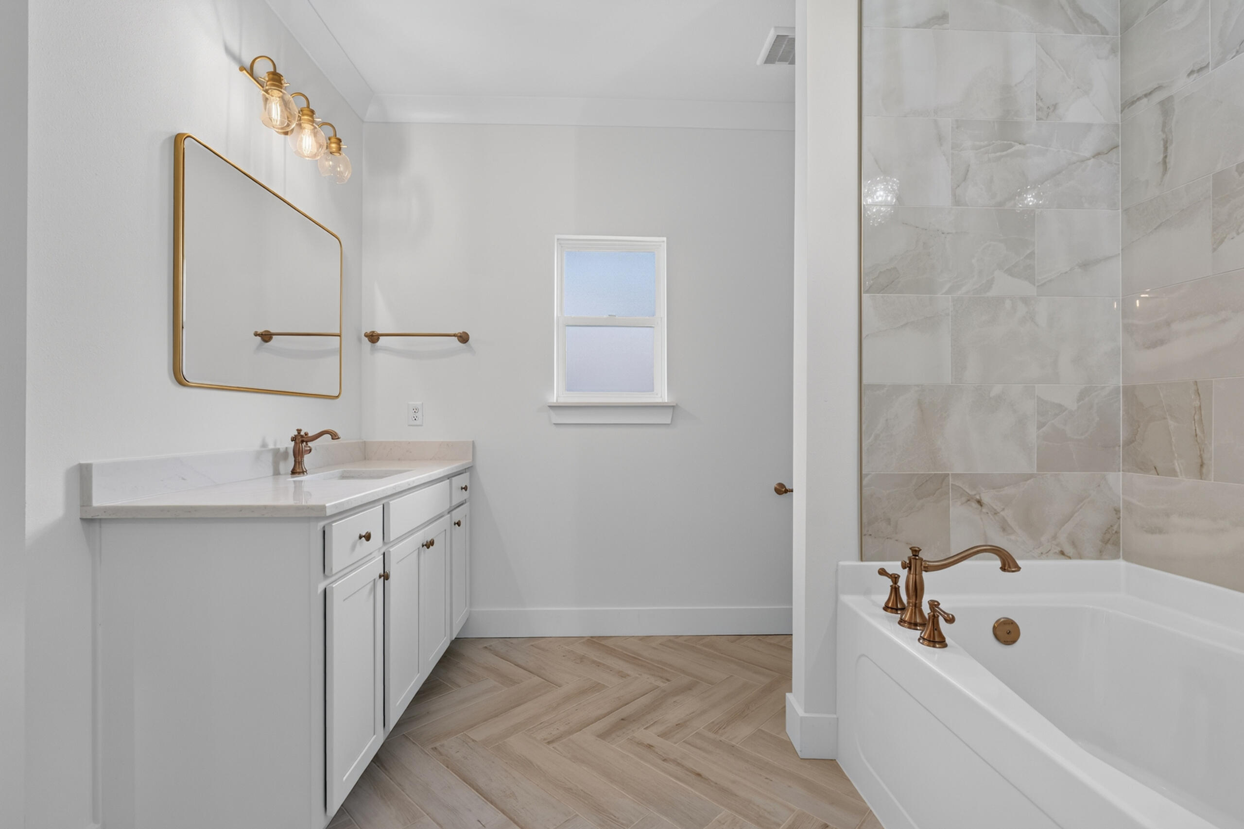 Bathroom with brass fixtures, a white vanity, marble-look tub surround, and herringbone tile floor.