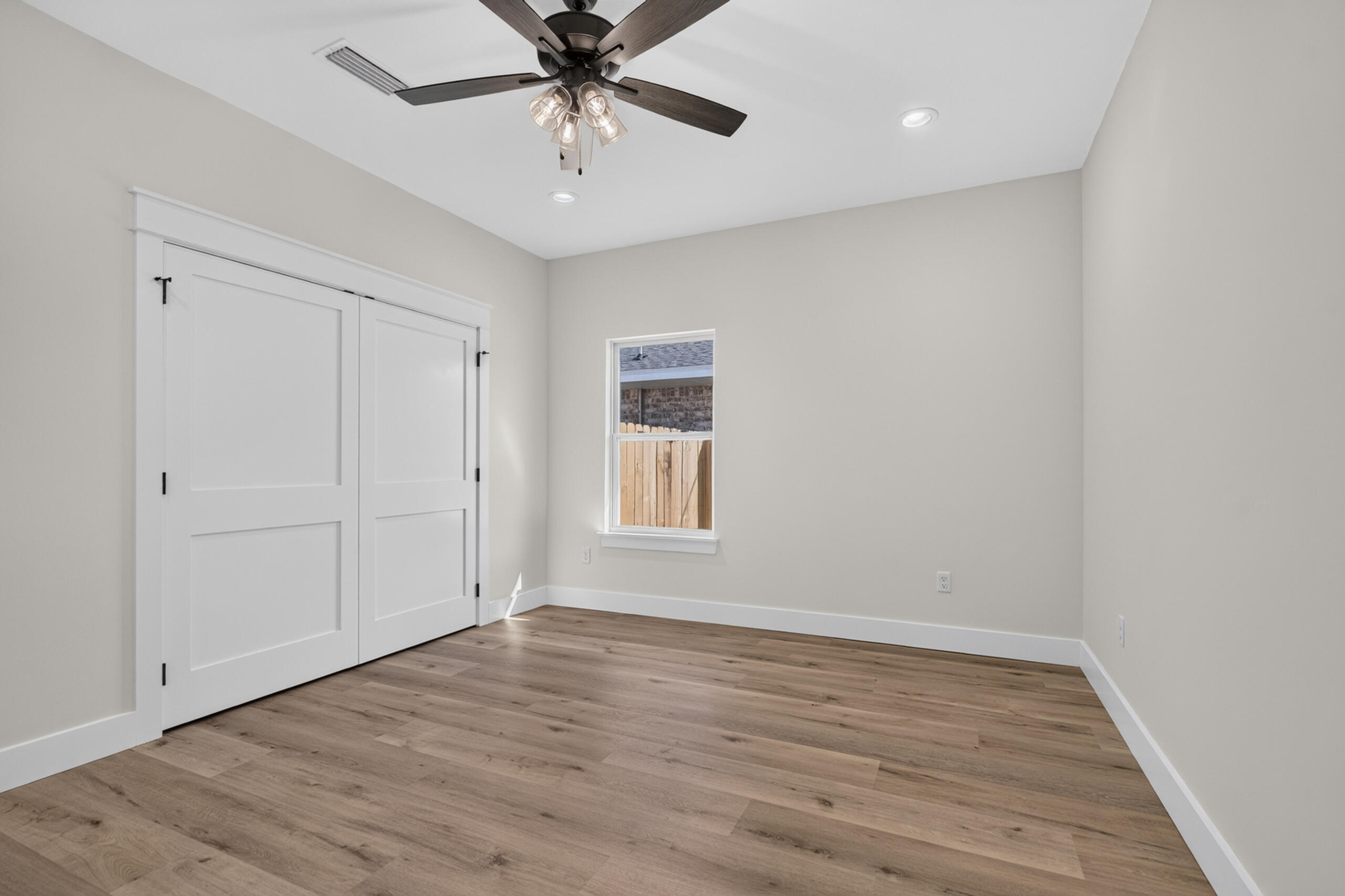 Empty bedroom with sliding white closet doors, wood-look floors, and a ceiling fan.