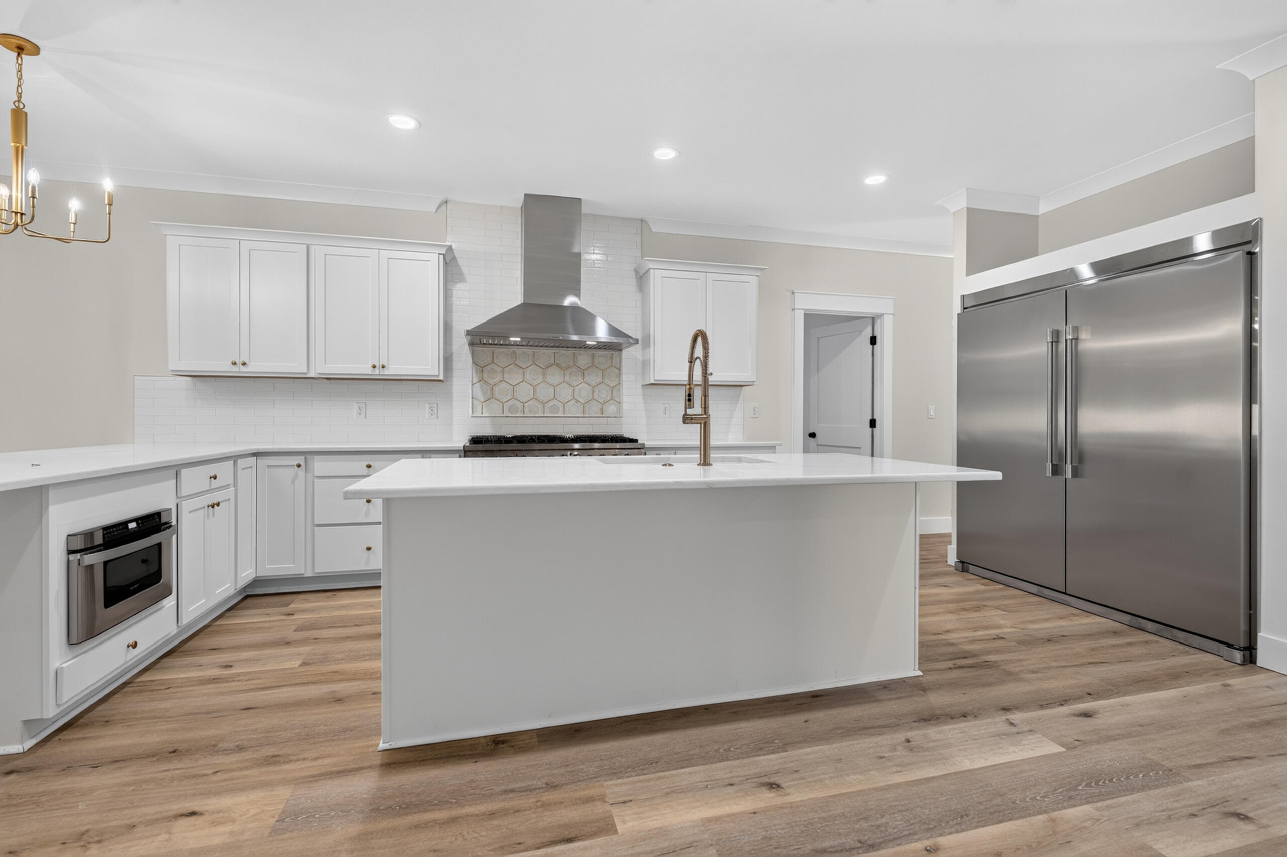 Modern white kitchen with a large island, brass faucet, stainless appliances, and wood-look flooring.