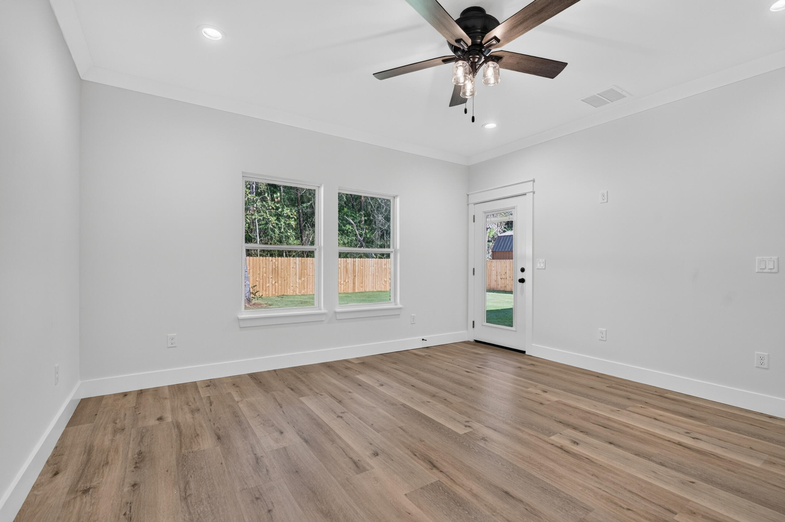 Vacant room with wood-look floors, ceiling fan, two windows, and a glass-panel door to a fenced yard.