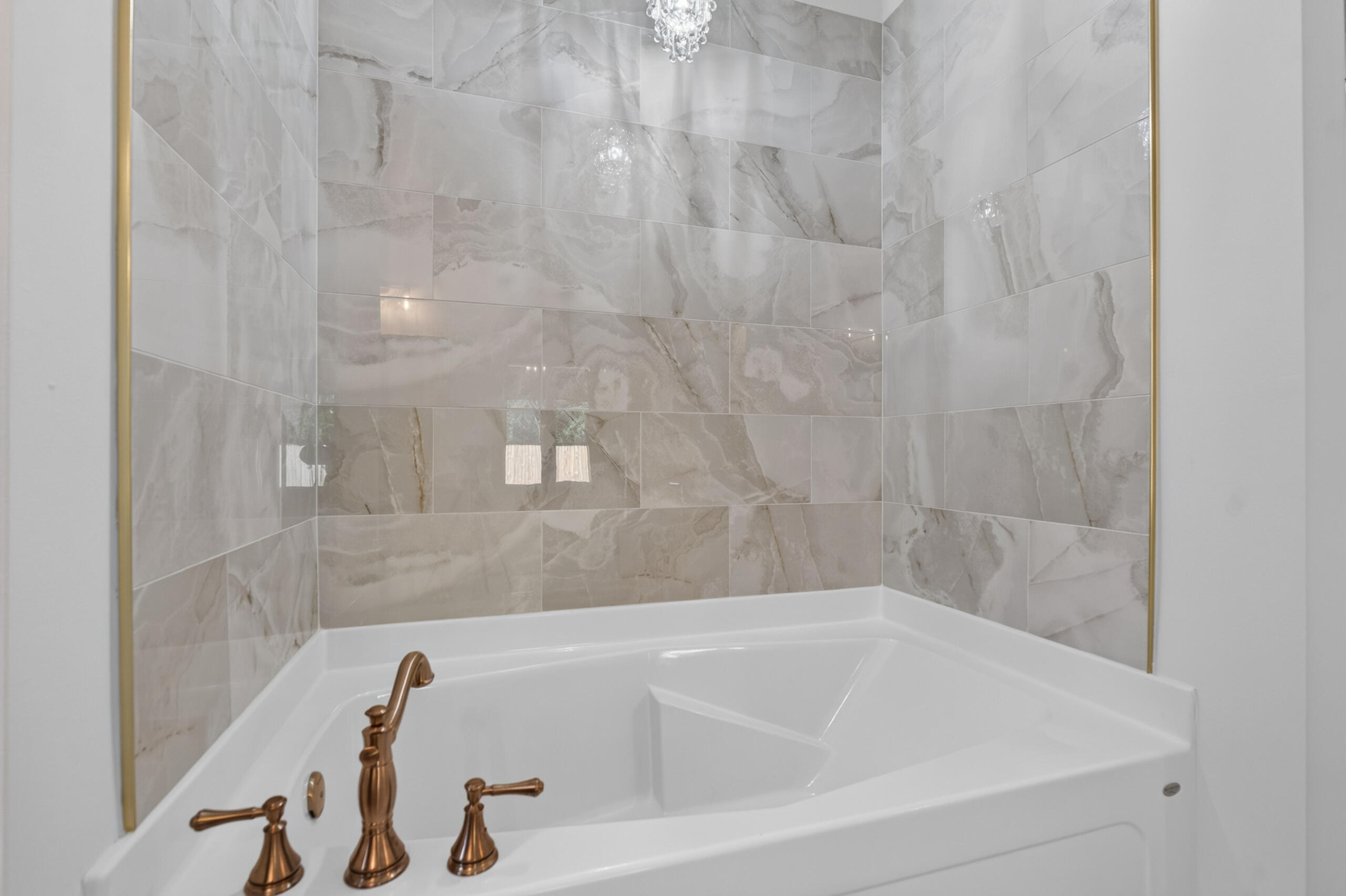 Corner bathtub with bronze fixtures, marble-look tile walls, gold trim, and a chandelier.