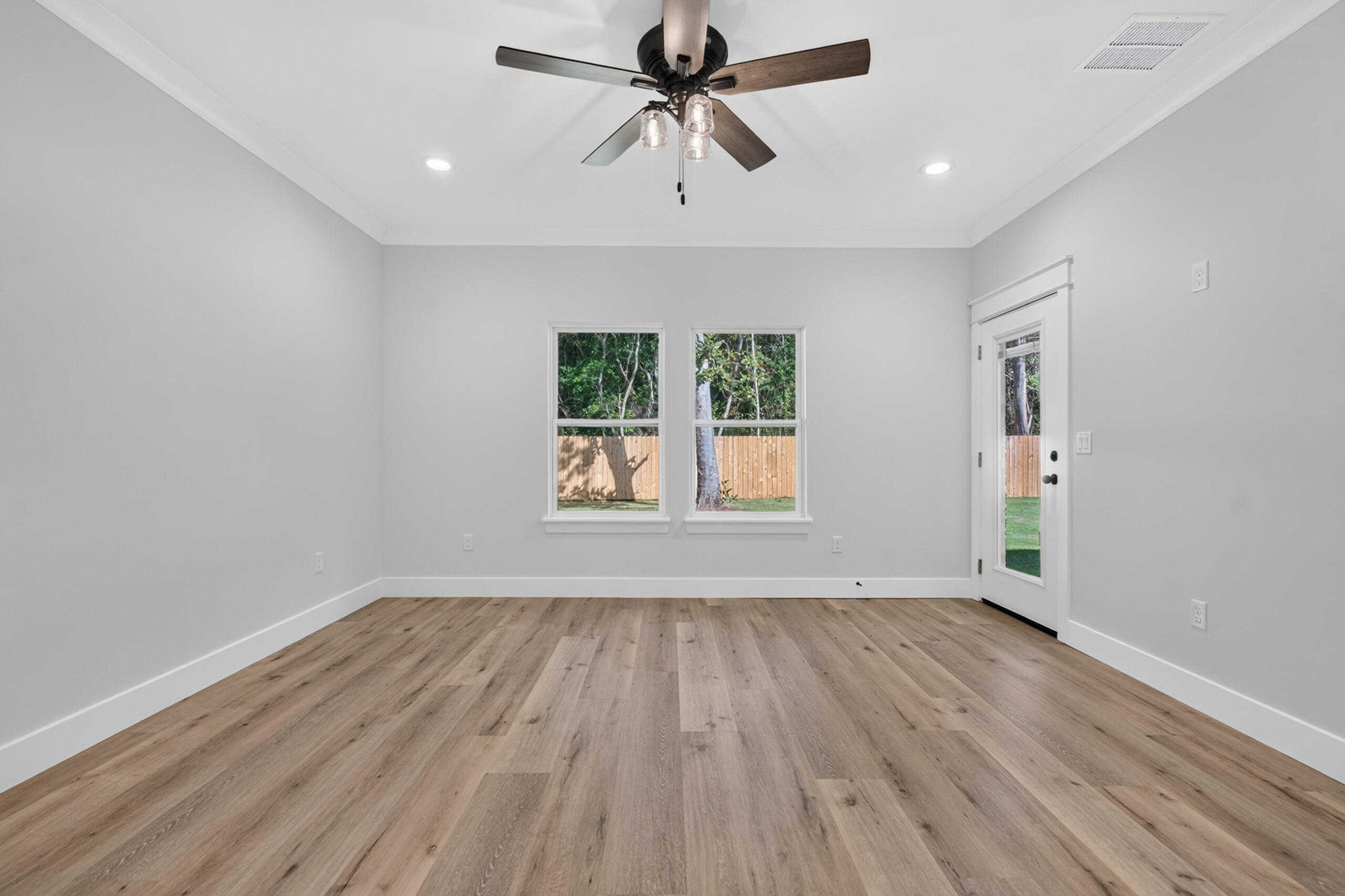 Empty room with wood-look flooring, ceiling fan, two windows, and a glass-paneled door to the yard.