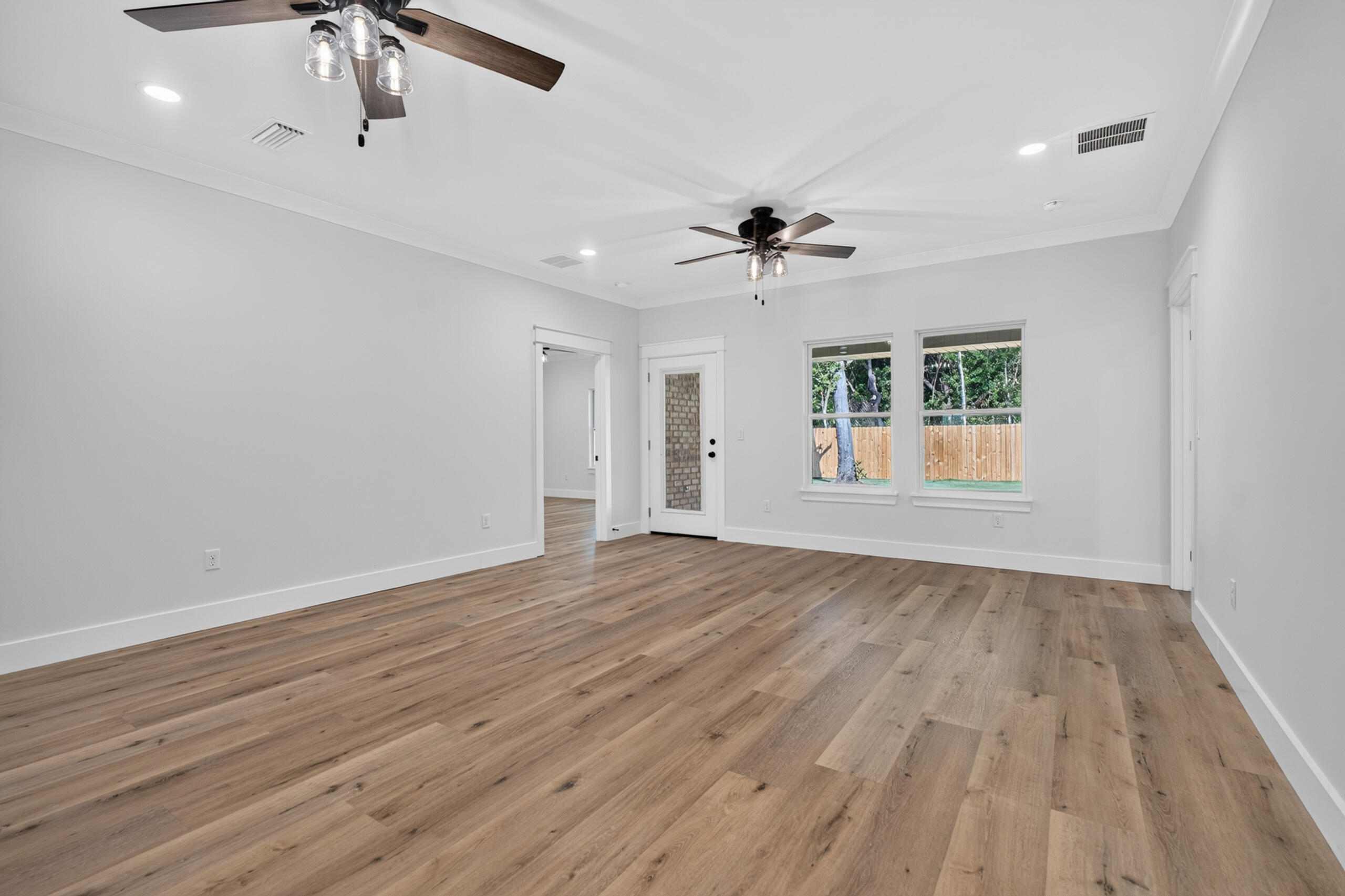 Vacant room with wood-look flooring, two ceiling fans, and windows facing a fenced yard.