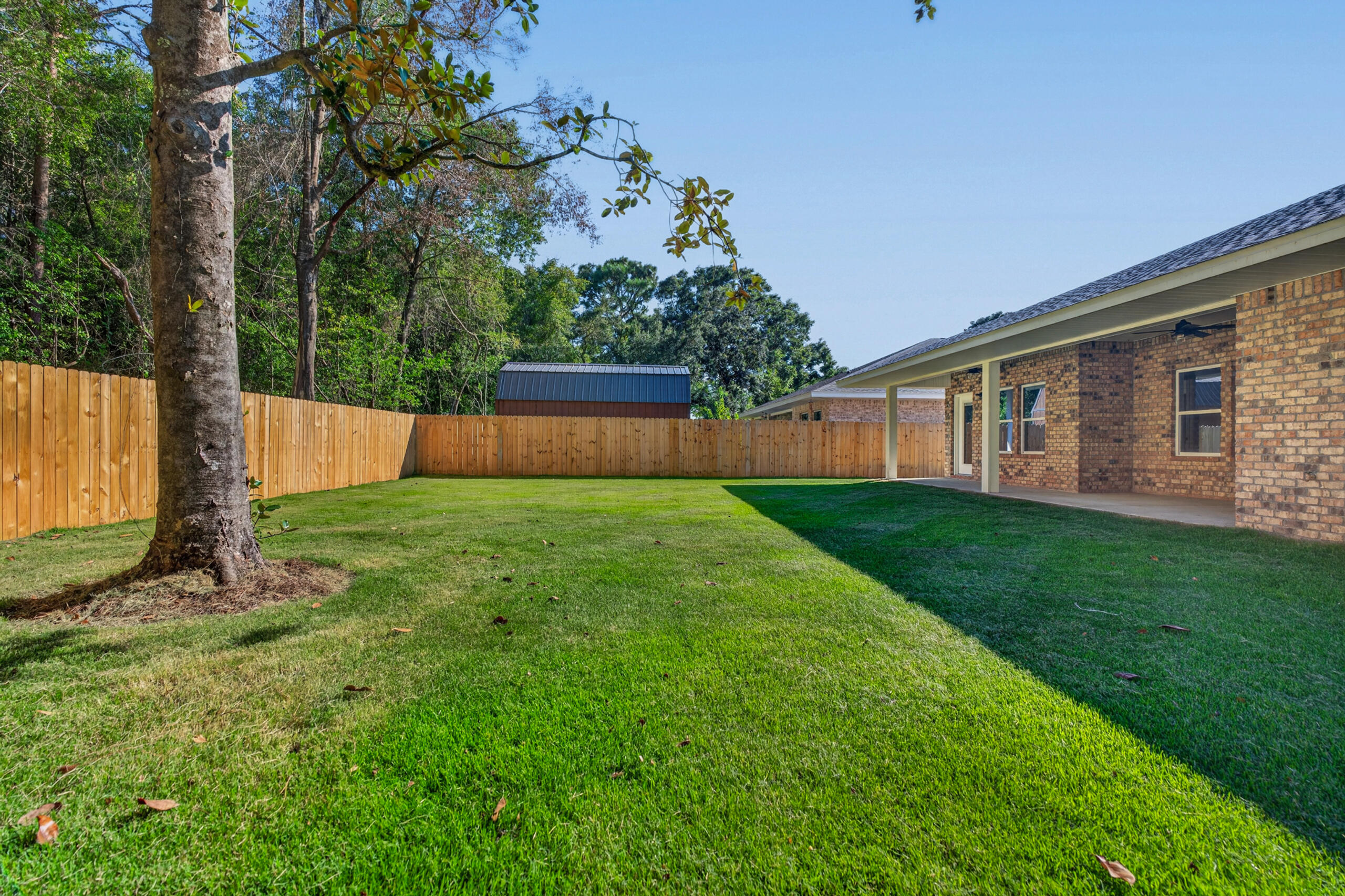 Fenced backyard with a broad lawn, mature tree, and covered patio beside a brick house.