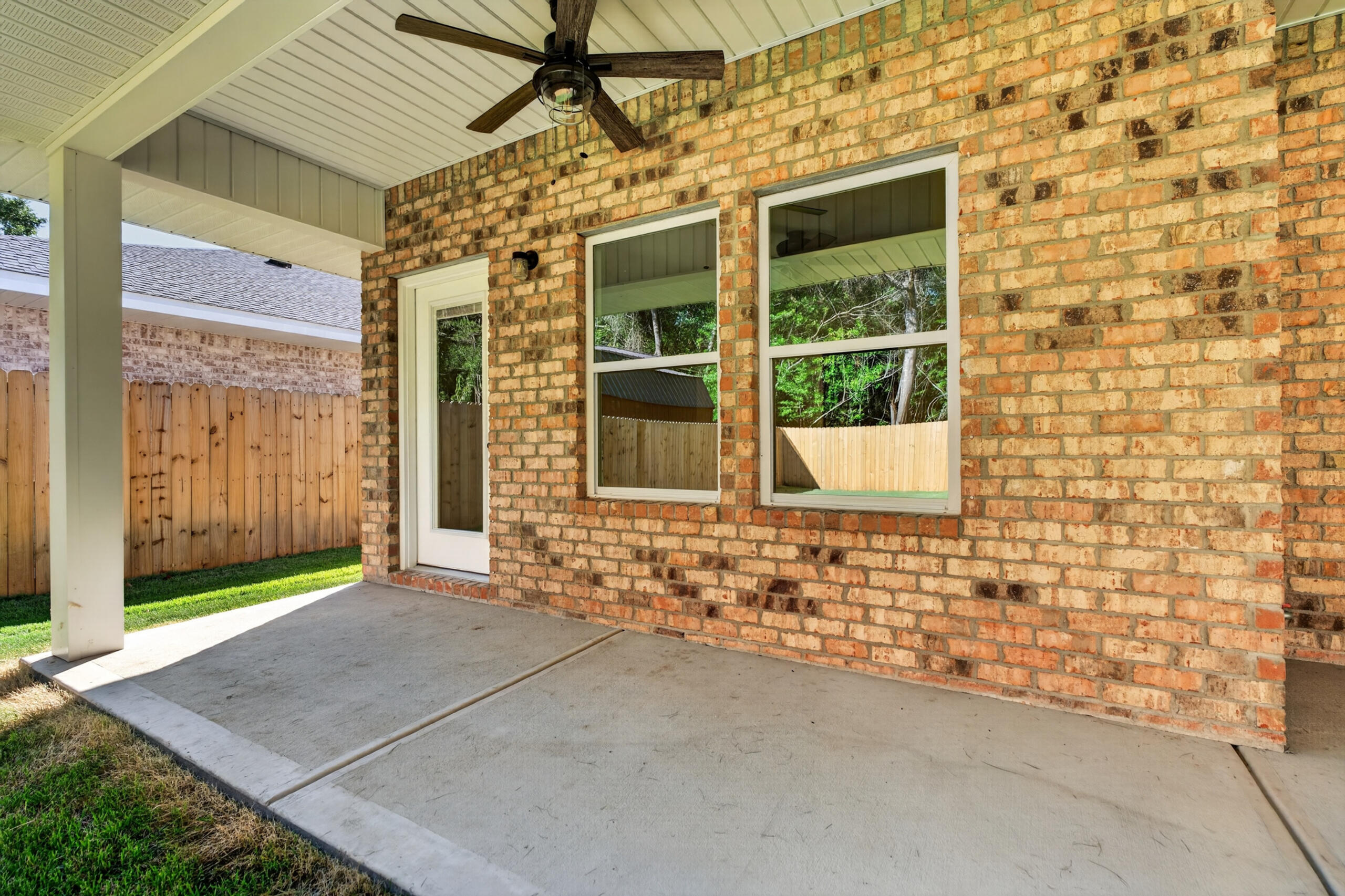 Covered concrete patio with a ceiling fan, brick wall, and windows overlooking a fenced yard.