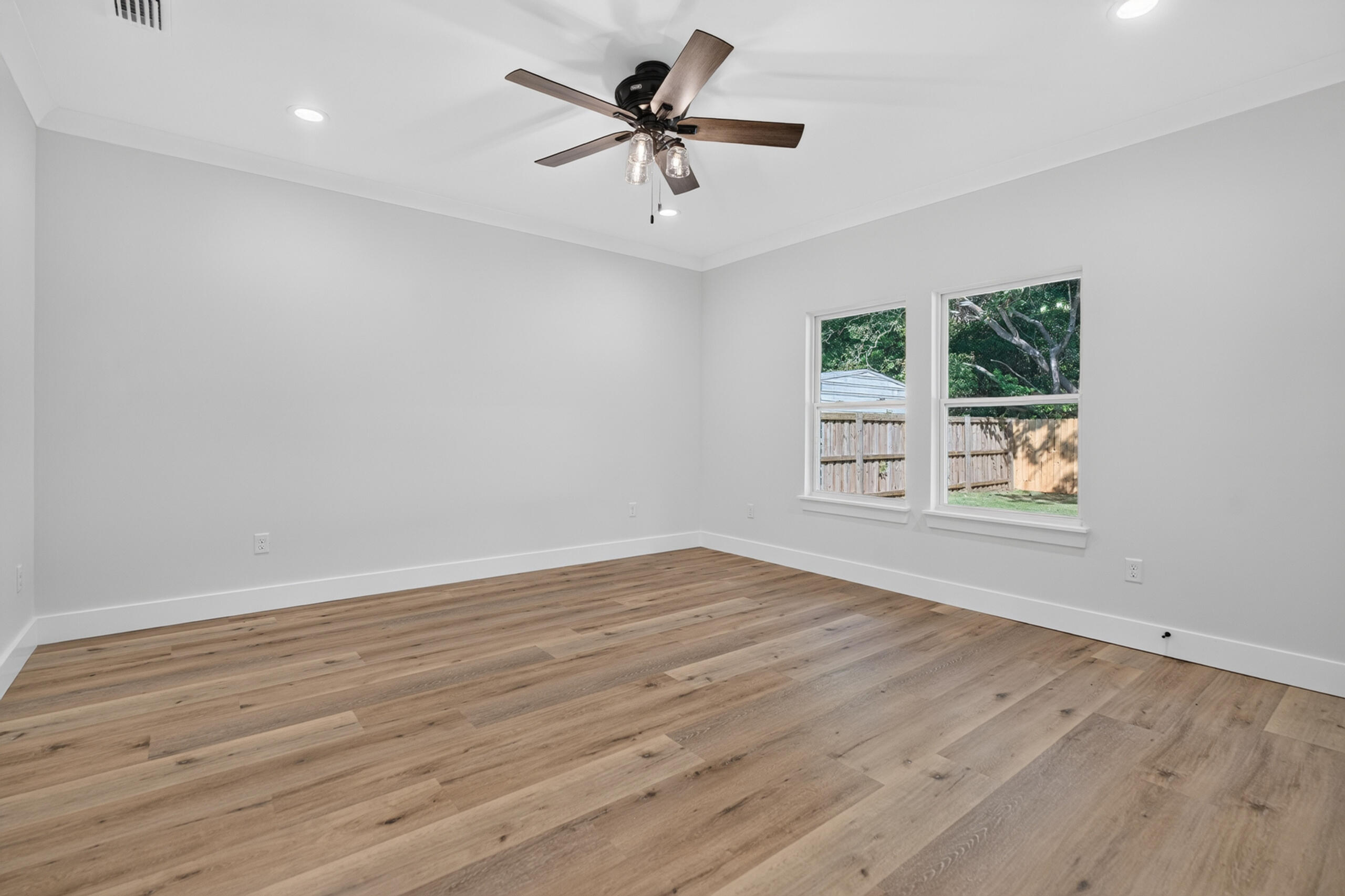 Empty bedroom with plank flooring, two yard-facing windows, and a dark-bladed ceiling fan.