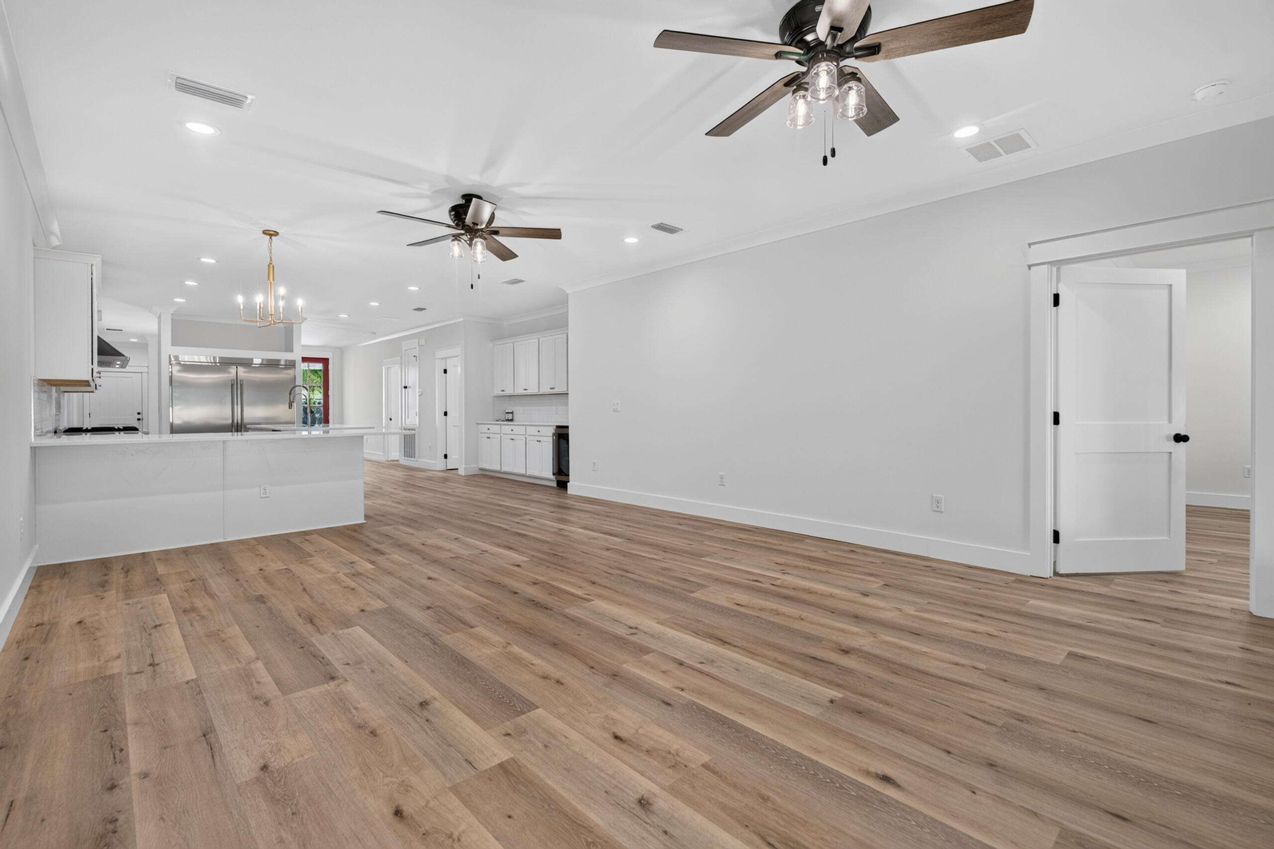 Empty living area with wood-look floors, adjoining a white kitchen with stainless steel appliances.