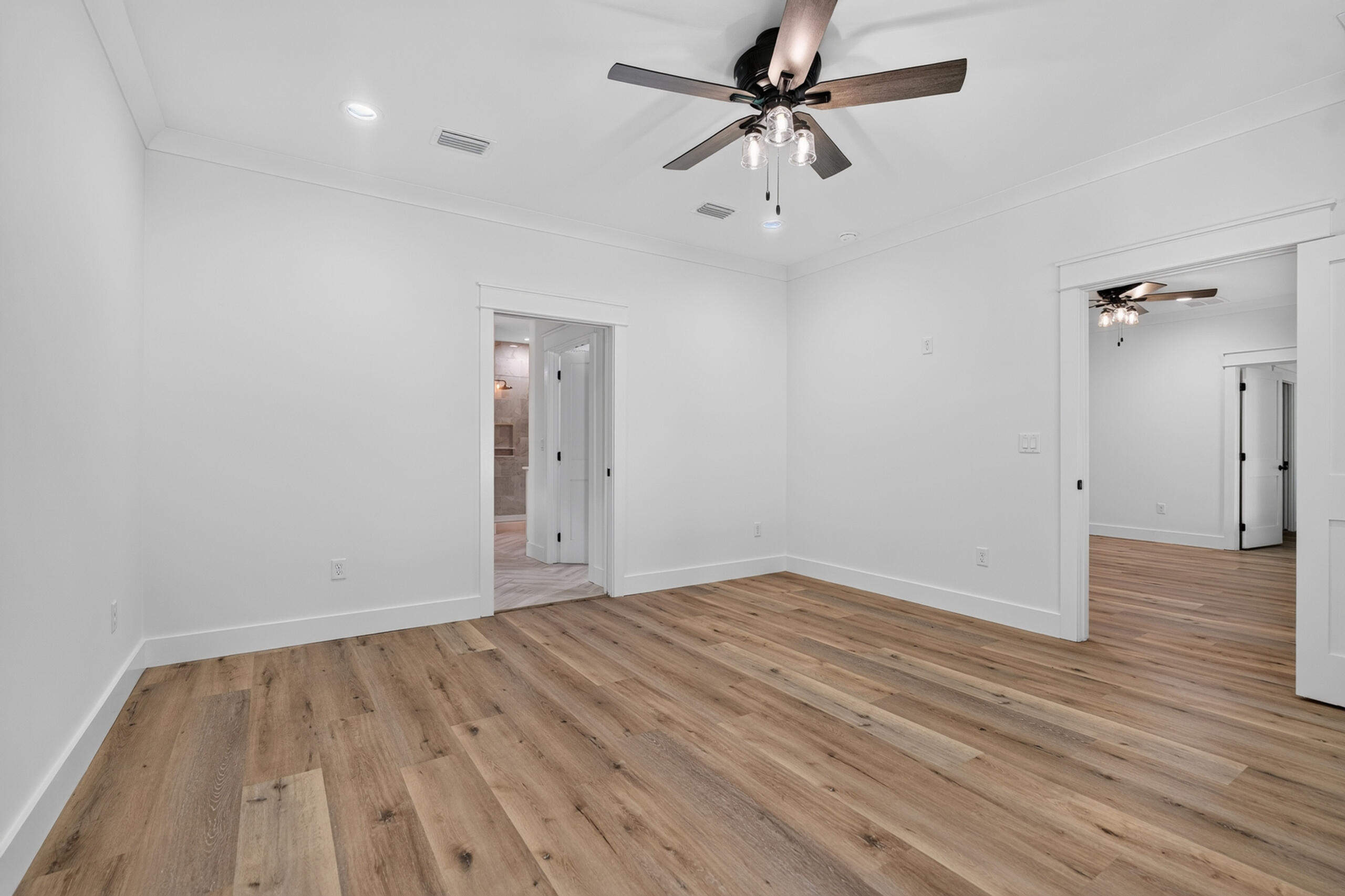 Empty bedroom with wood-look flooring, a ceiling fan, and open doorways to adjoining rooms.