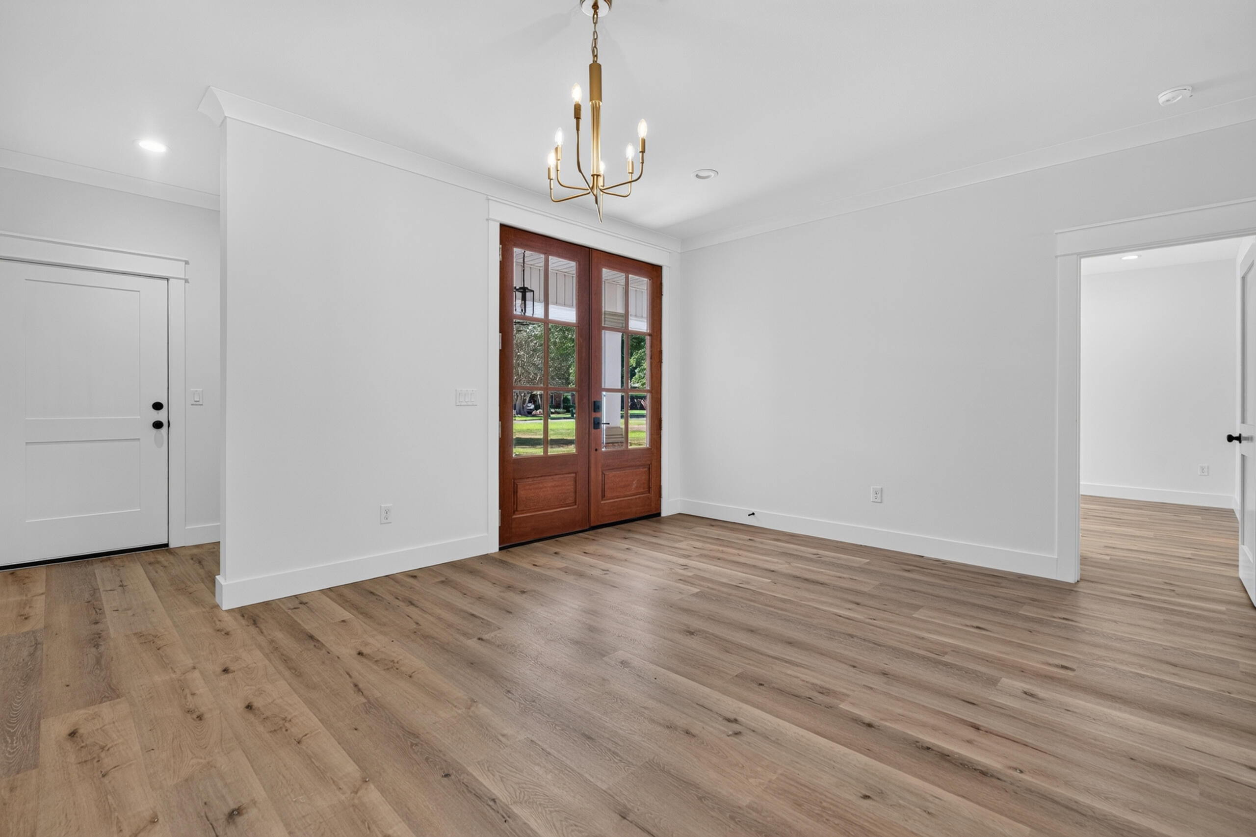 Empty room with light wood floors, brass chandelier, and glass-paneled French doors.