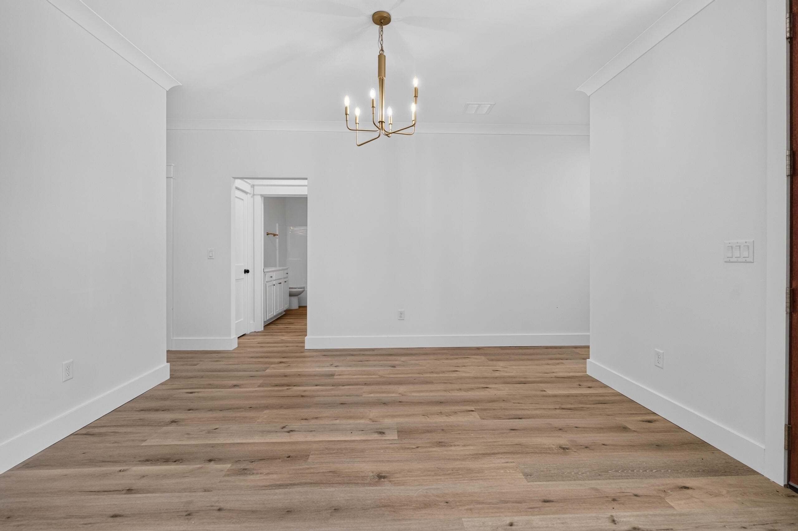 Empty room with wood-plank flooring and a brass chandelier, opening to a bathroom.