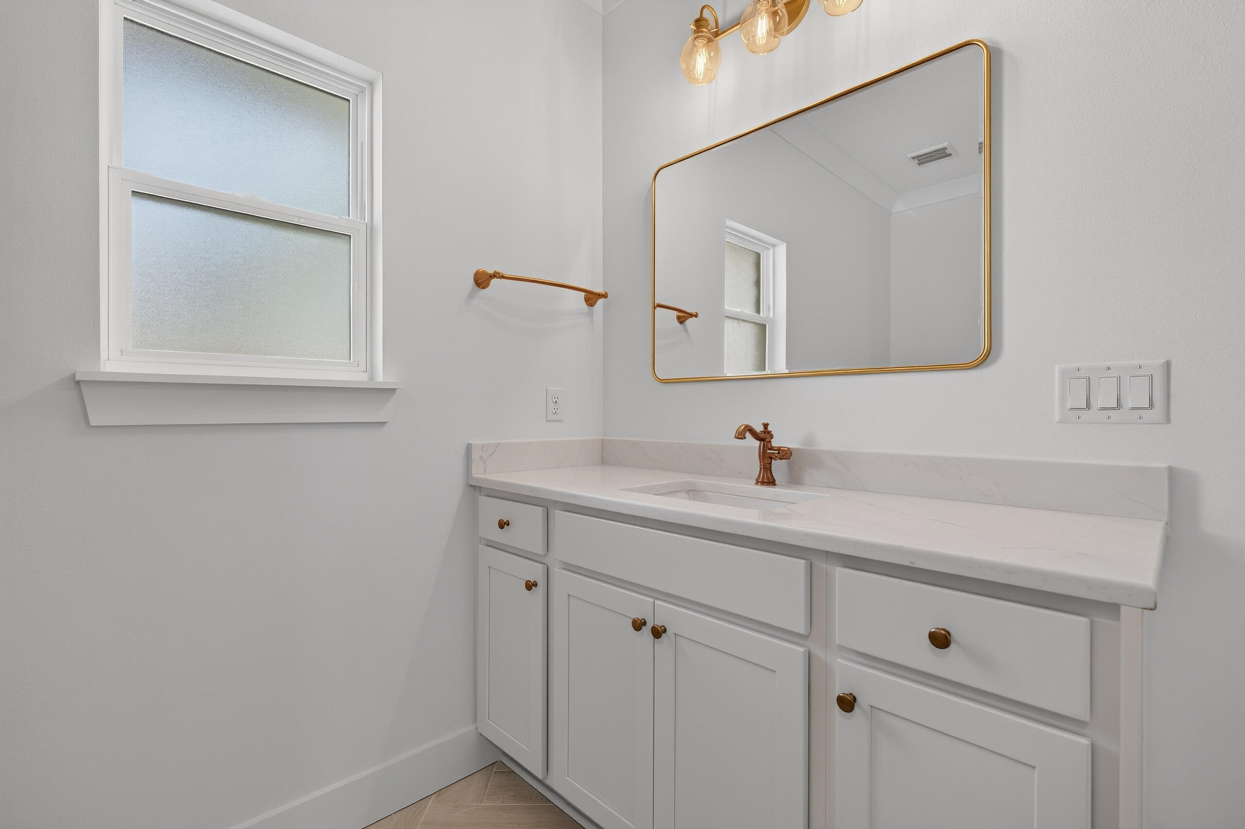 Bathroom vanity with white cabinetry, marble-look counters, brass fixtures, and a gold-framed mirror.