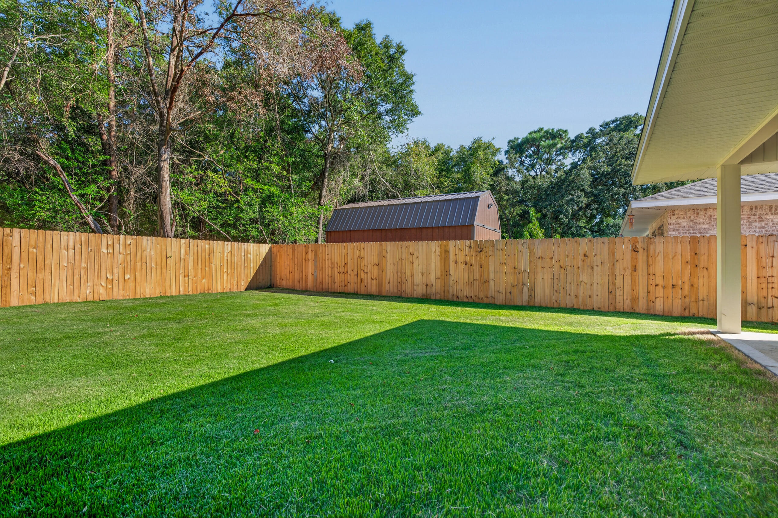 Fenced backyard with a broad green lawn, covered patio edge, and shed beyond the fence.