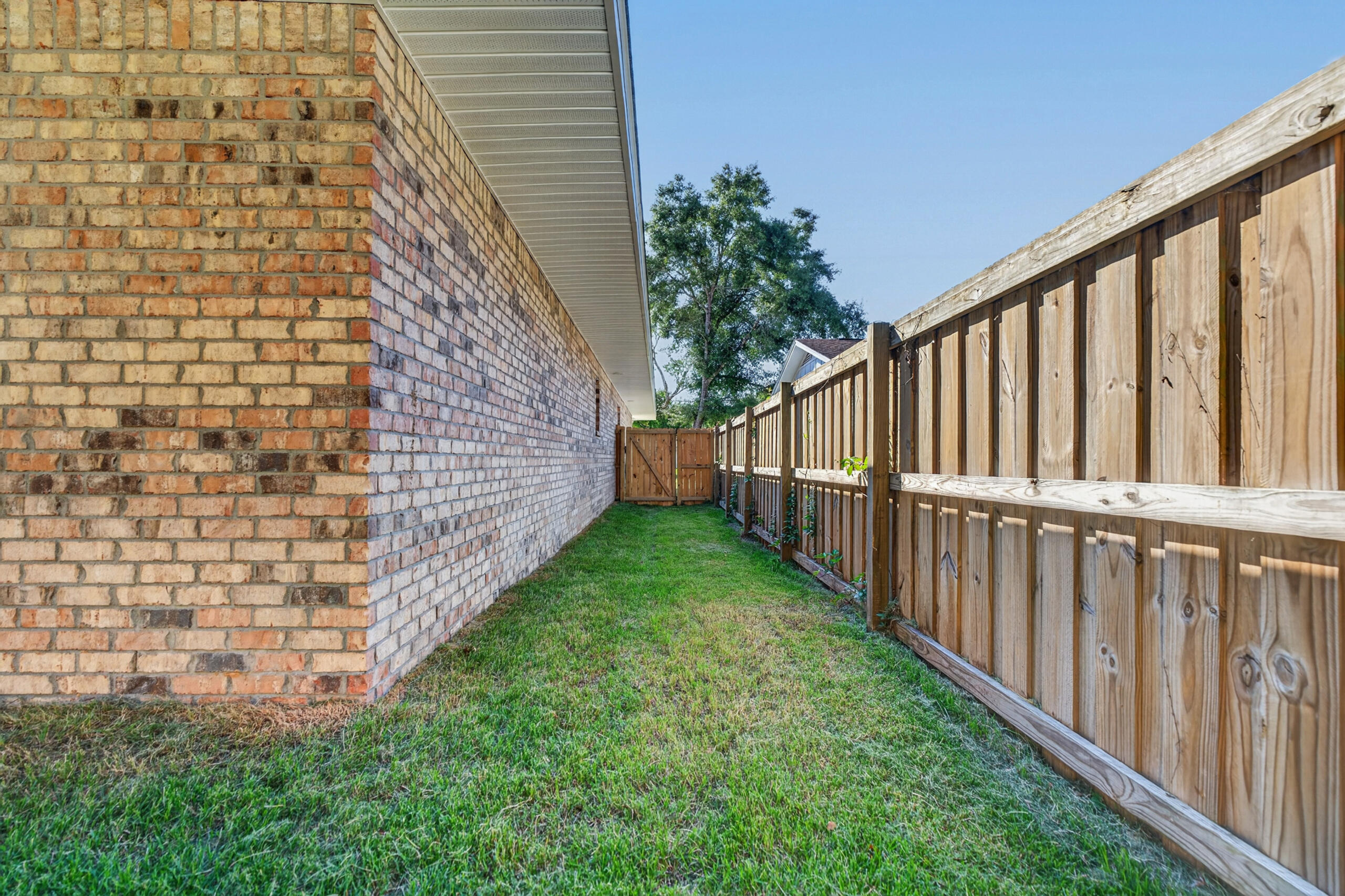 Grassy side yard between a brick house and wooden fence, with a gate at the far end.