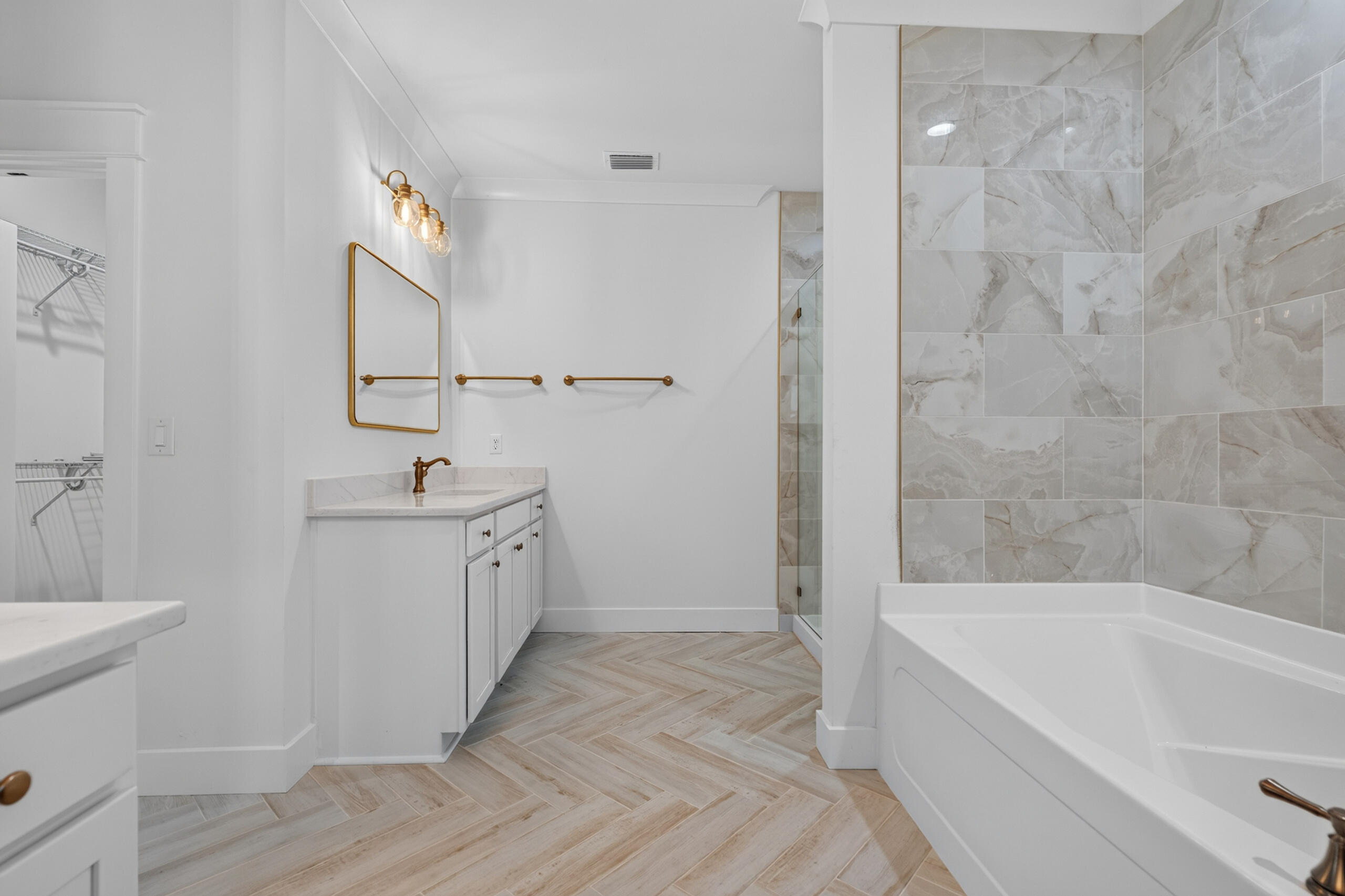Bathroom with a brass-accented vanity, glass shower, white tub, and herringbone tile floor.