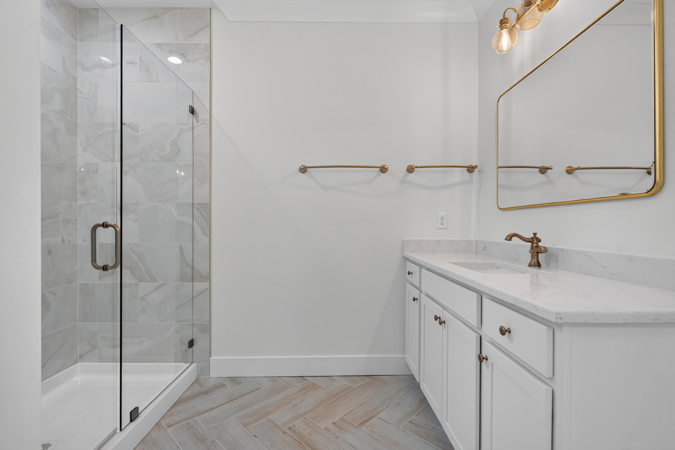 Bathroom with a glass shower, white vanity, gold-tone fixtures, and light herringbone tile floor.