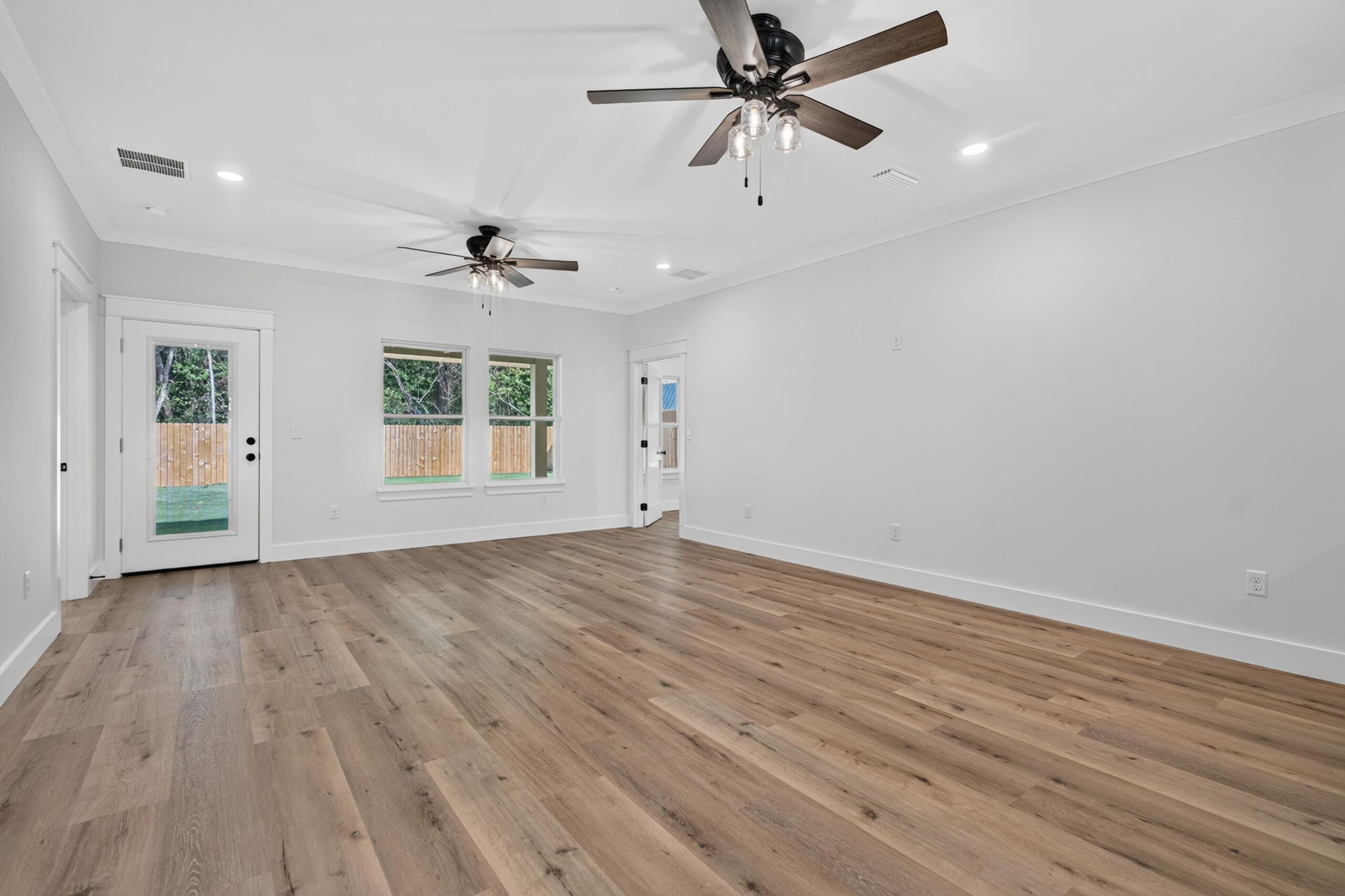 Empty living room with wide-plank wood flooring, two ceiling fans, and windows facing a fenced yard.
