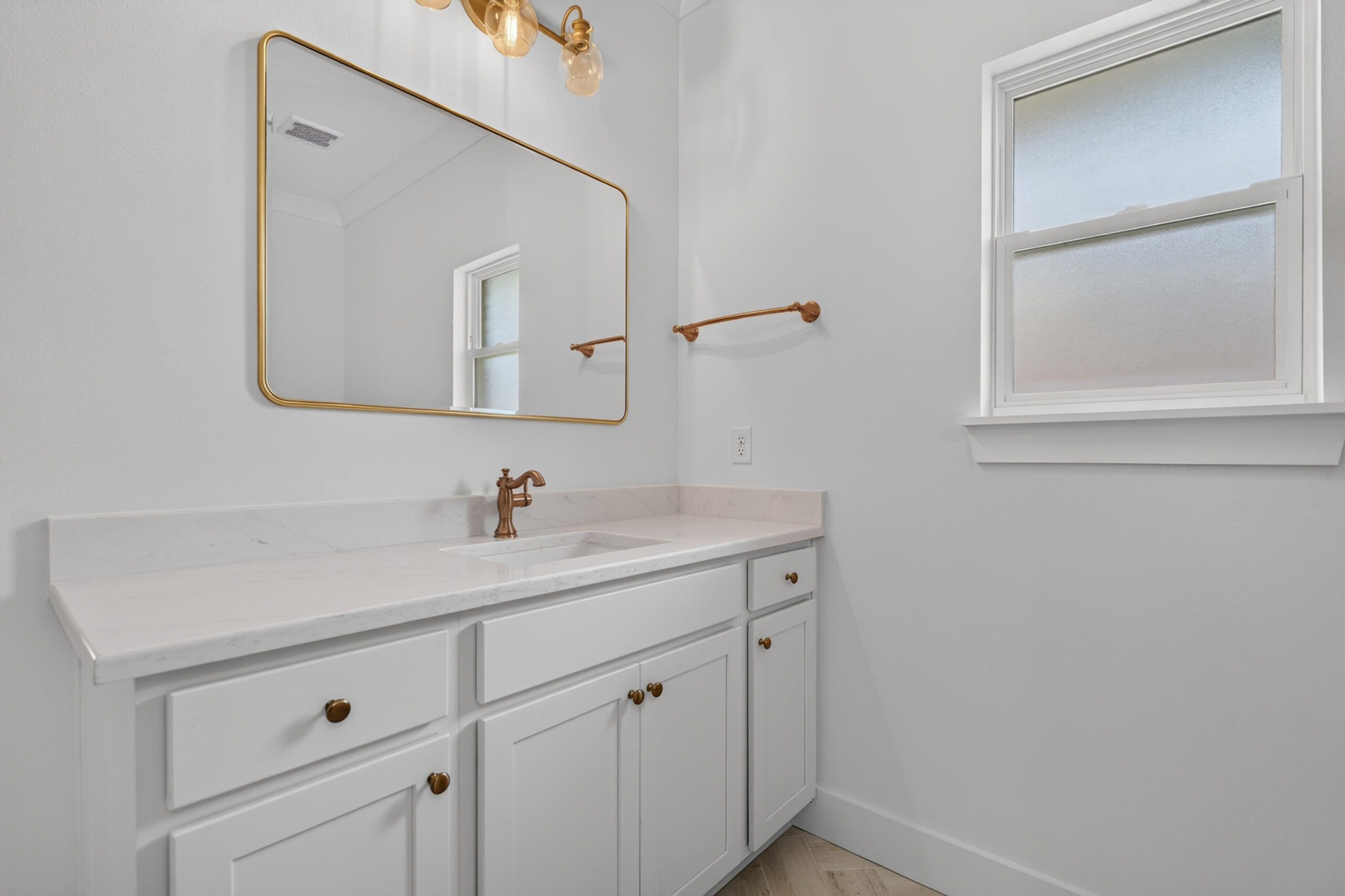 Bathroom vanity with white cabinets, brass fixtures, a gold-framed mirror, and a frosted window.