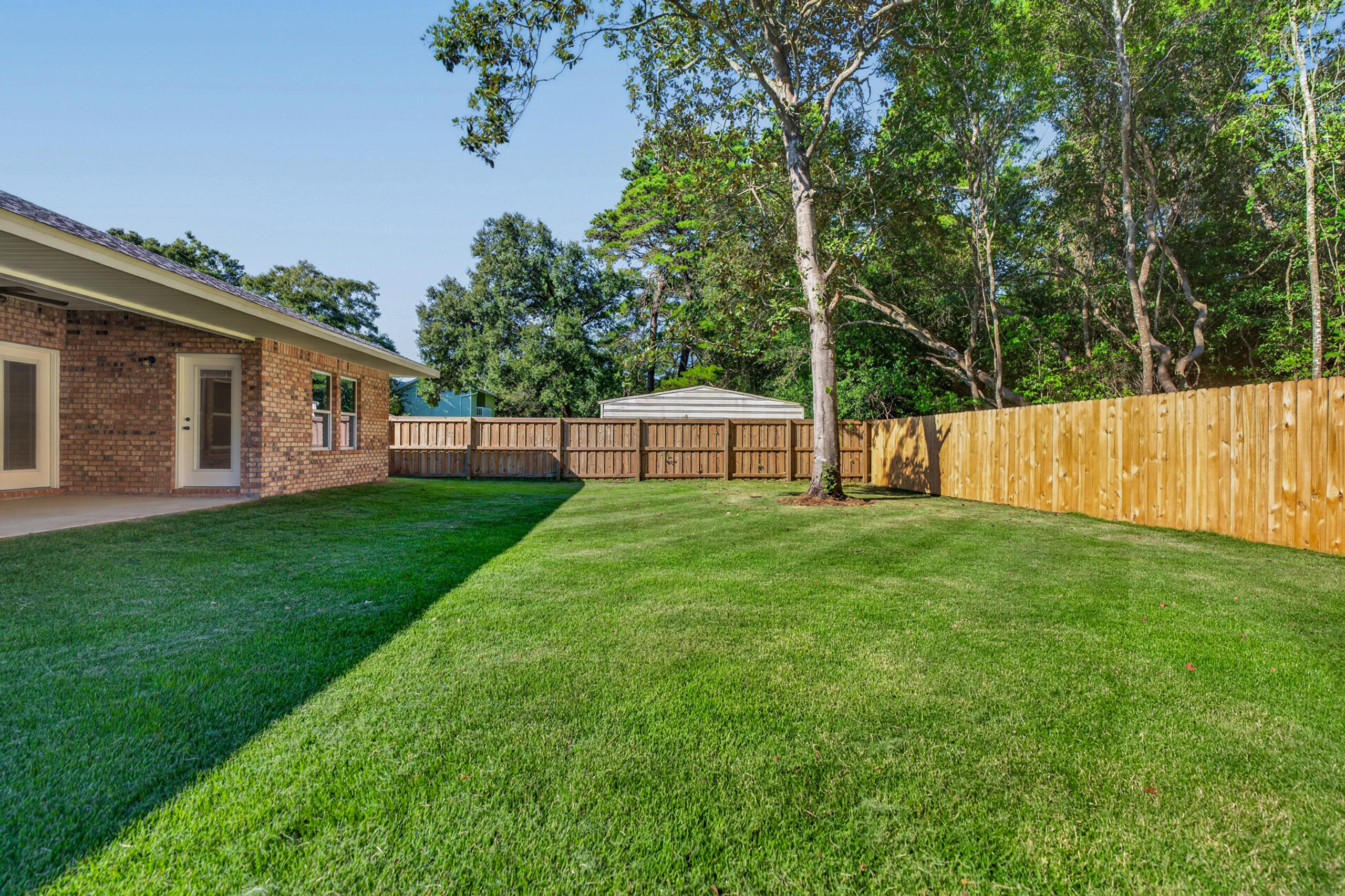 Fenced backyard with a broad lawn, mature trees, and a brick home.
