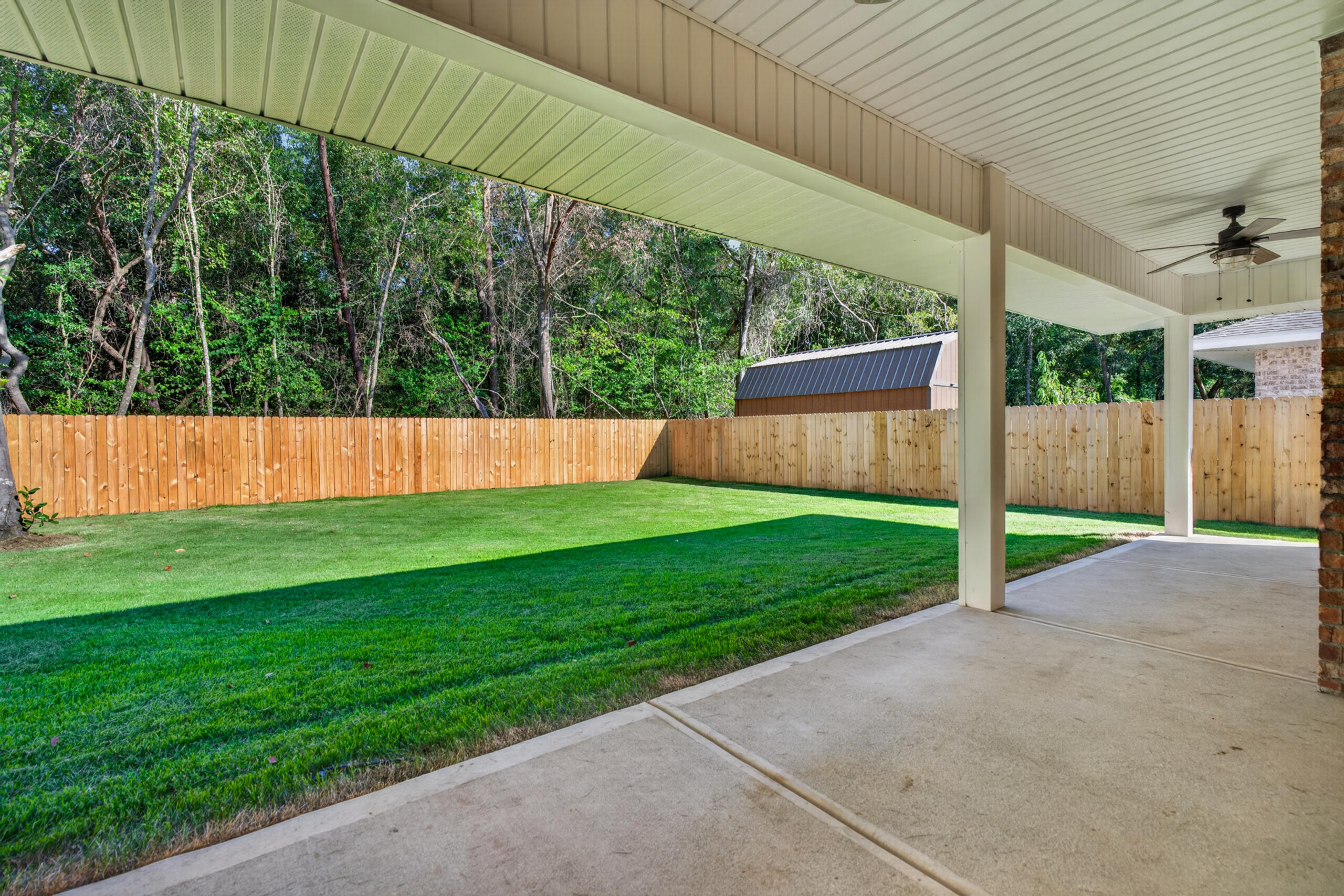Covered patio overlooking a fenced, grassy backyard with mature trees and a small shed.