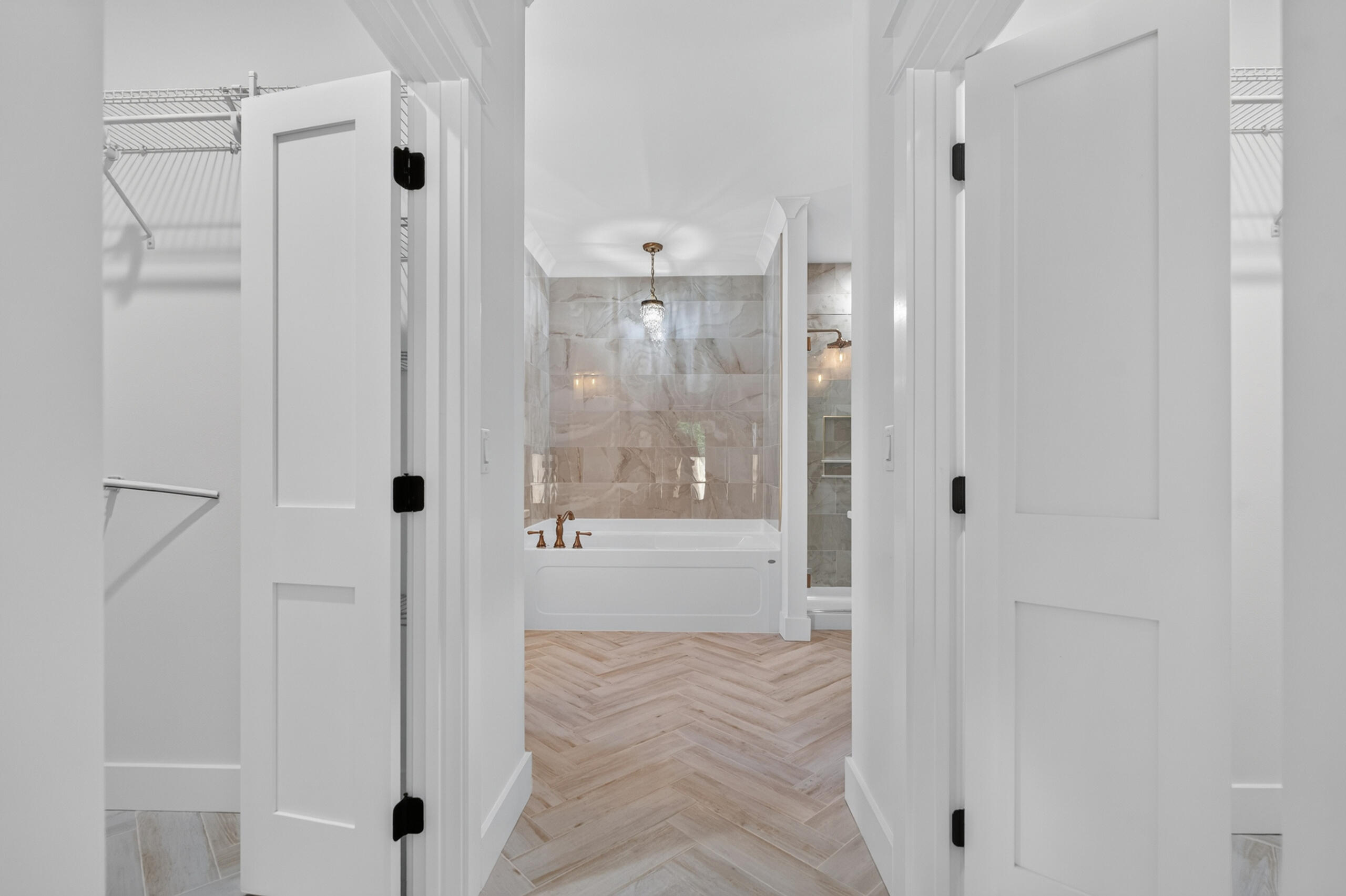 Bathroom with a marble-look tiled tub surround, crystal pendant, and light herringbone flooring.