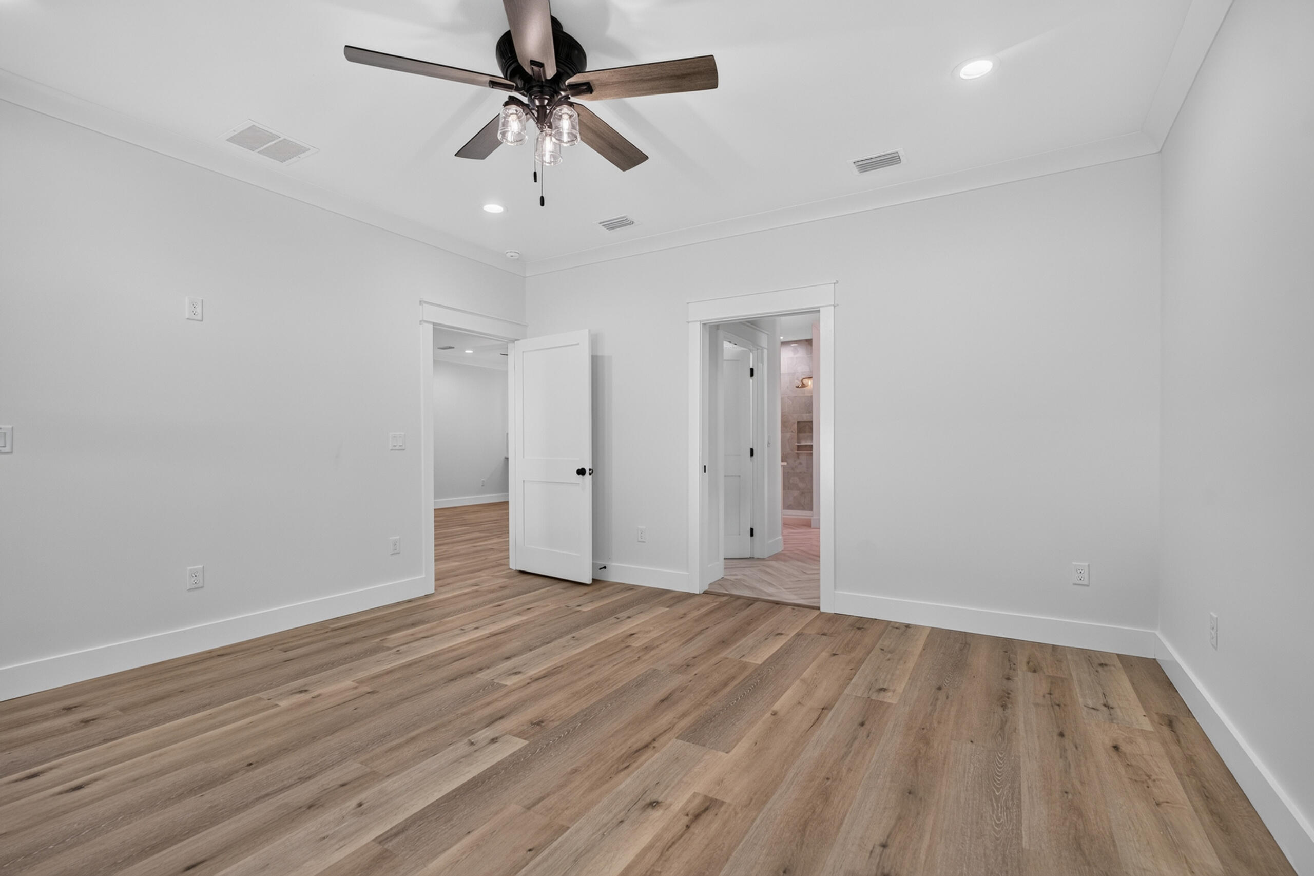 Empty bedroom with wide-plank wood floors, a dark ceiling fan, and an open doorway to a tiled bathroom.