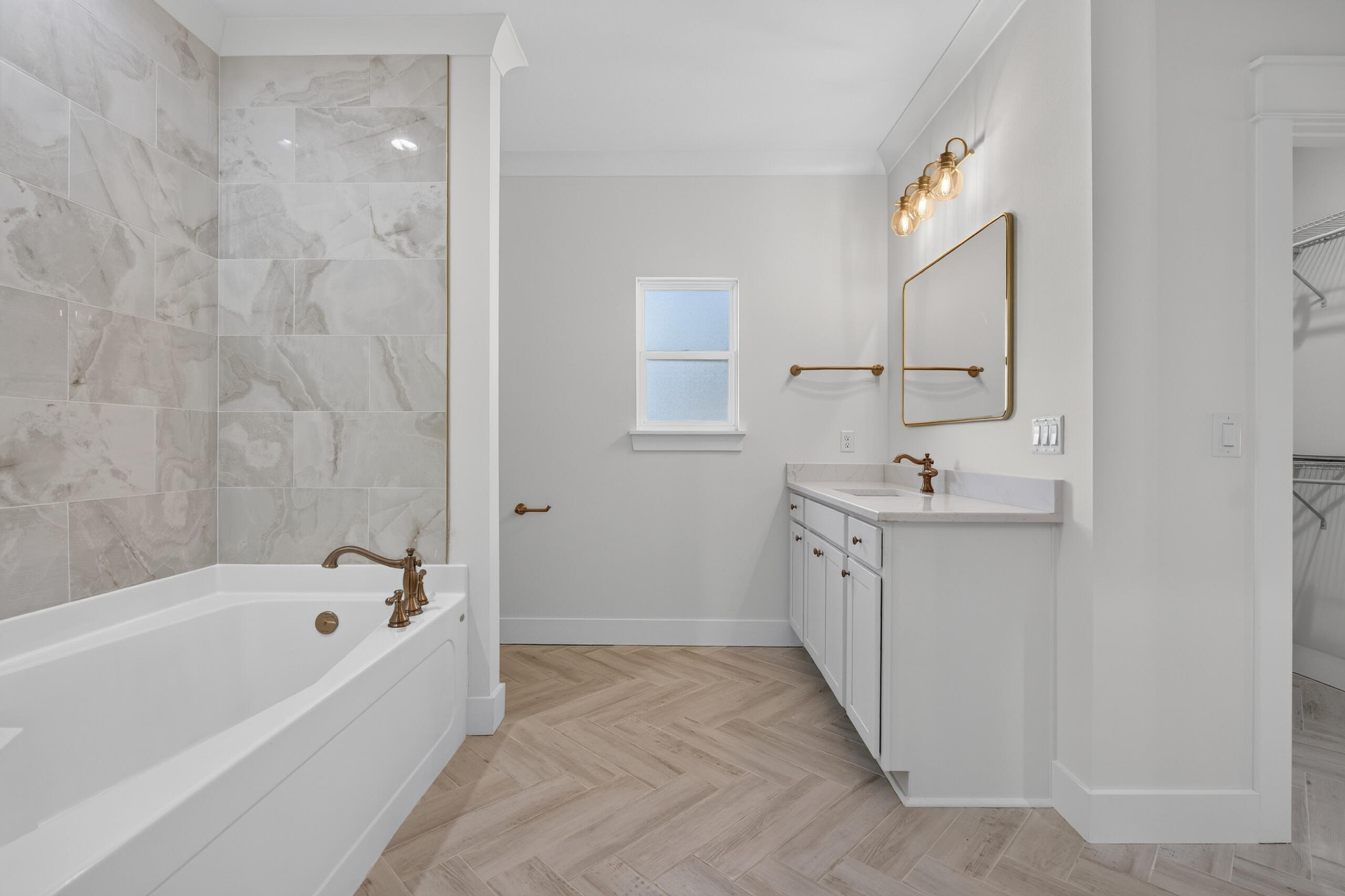 Bathroom with a marble-look tiled tub surround, brass fixtures, white vanity, and herringbone floor.