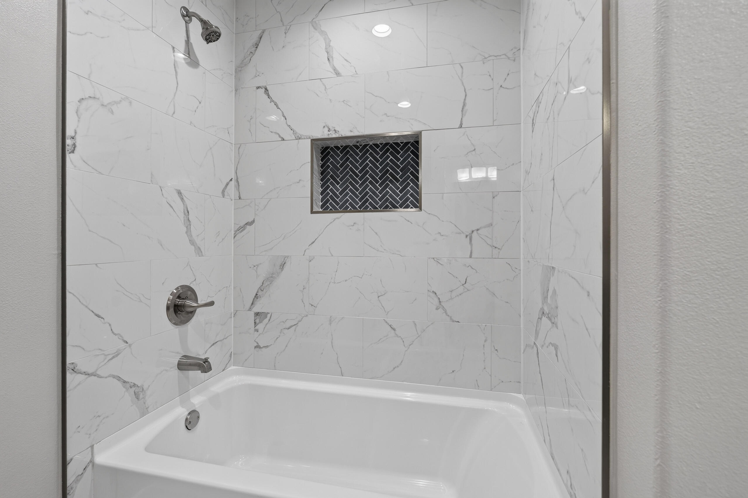 Bathtub and shower with marble-look tile and a dark herringbone niche.