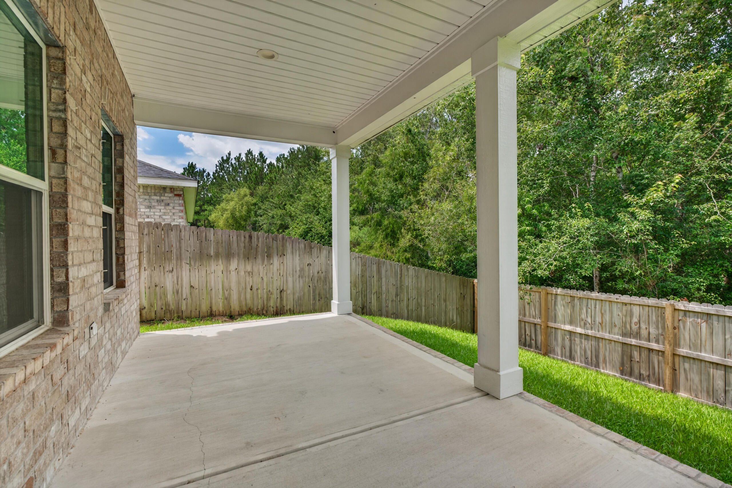 Covered concrete patio beside a brick house, overlooking a fenced yard and leafy trees.