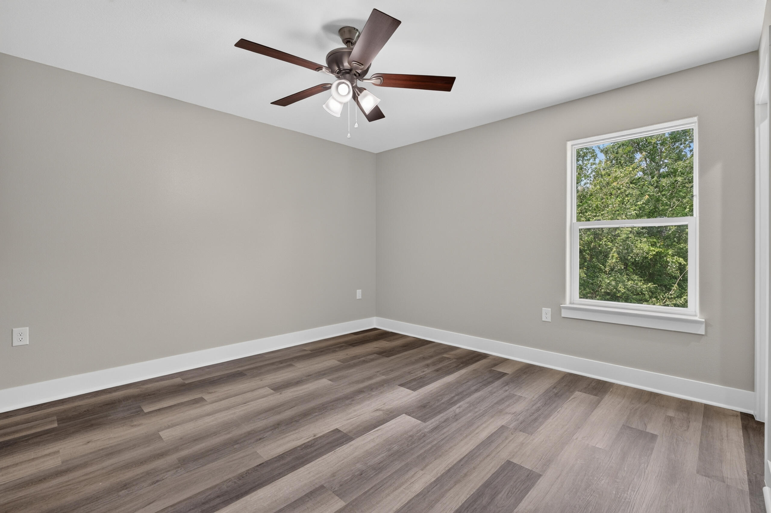 Empty bedroom with wood-look floors, a ceiling fan, and a window overlooking trees.