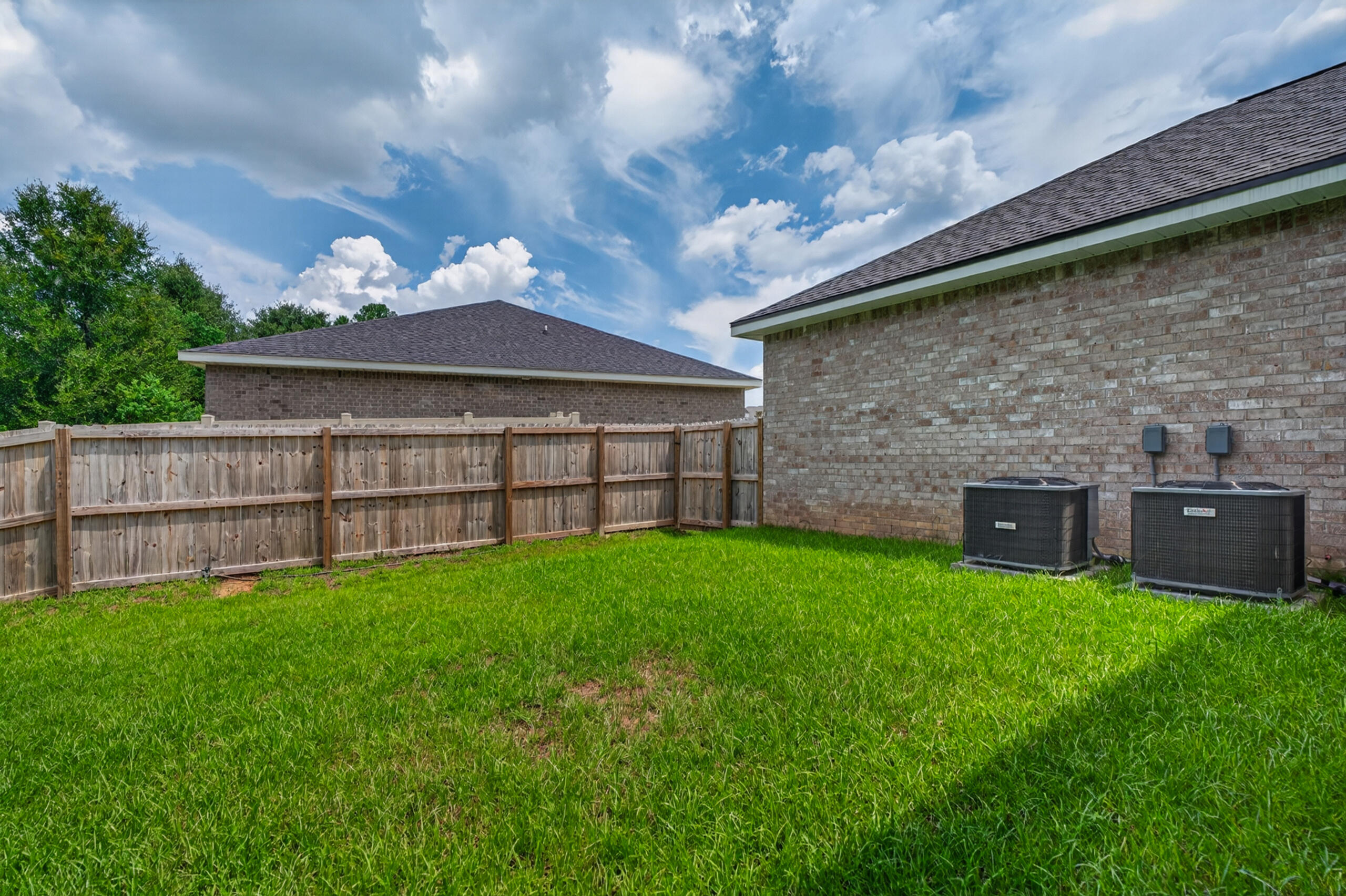 Fenced backyard with a green lawn and two air-conditioning units beside a brick house.
