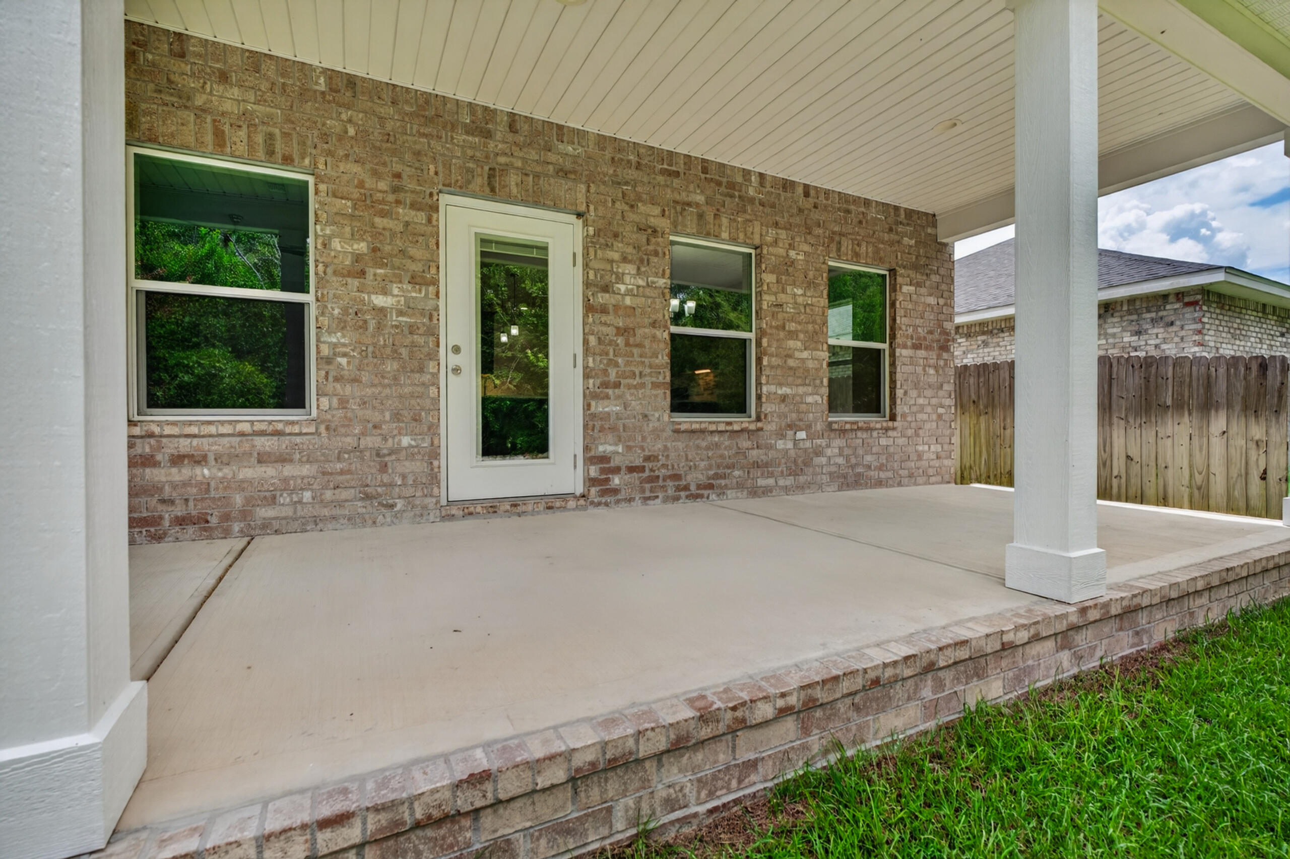 Covered concrete porch against a brick wall, with white columns, a glass door, and a low brick step.
