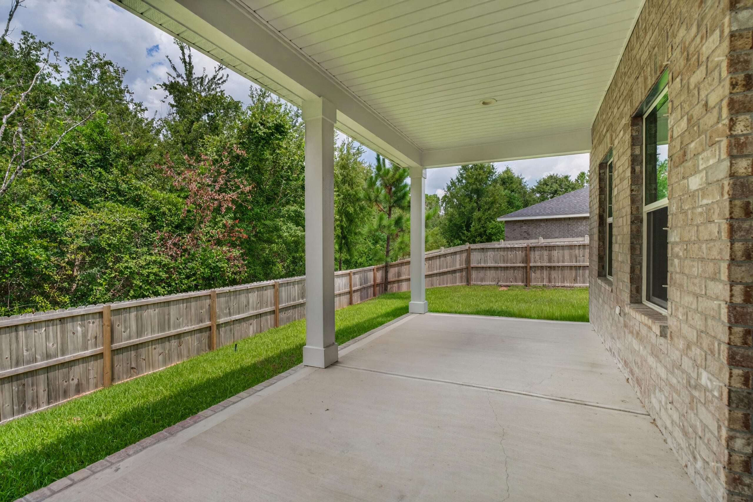 Covered concrete patio beside a brick home, overlooking a fenced grassy yard and mature trees.