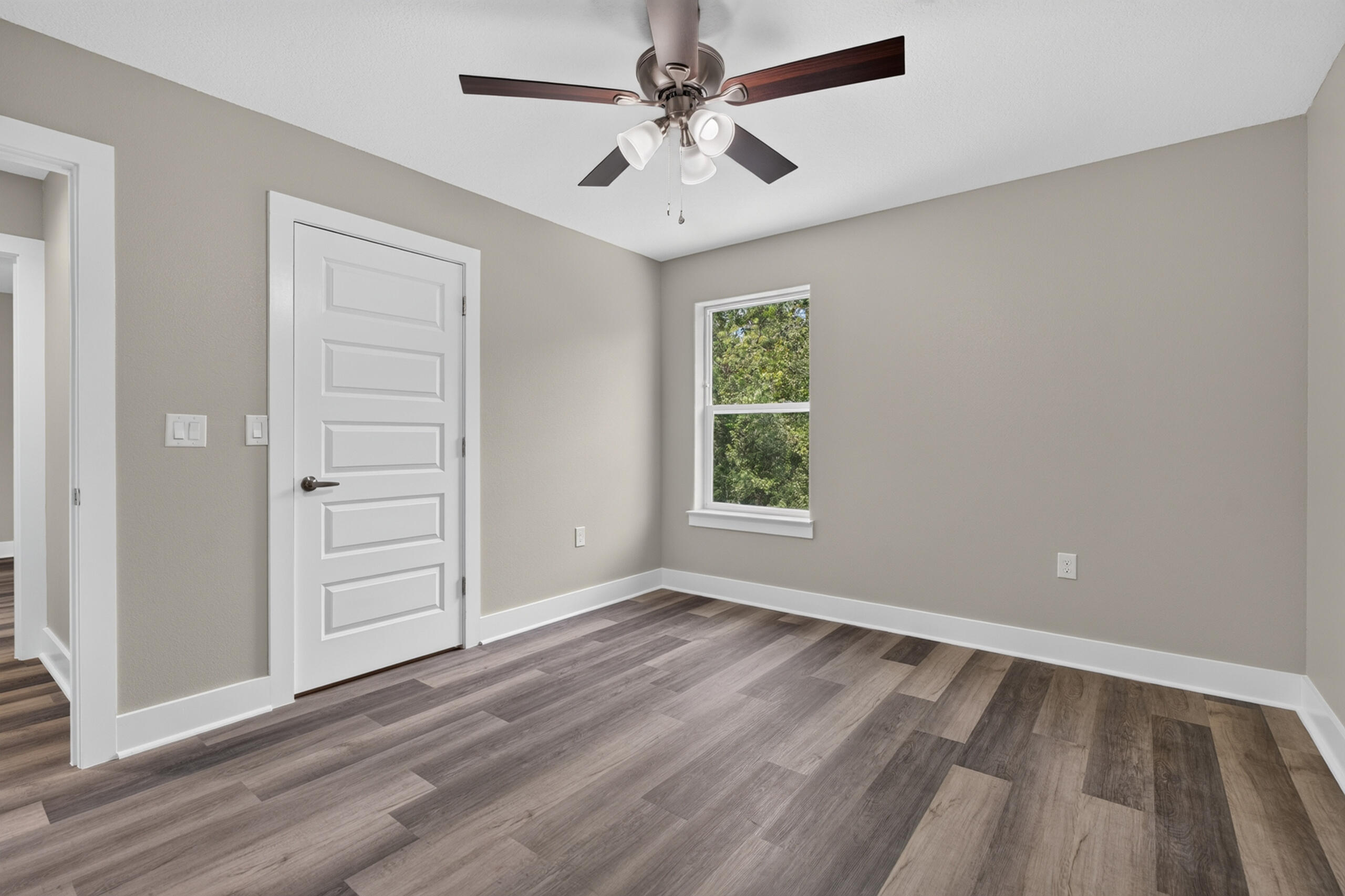 Empty bedroom with wood-look flooring, a tree-facing window, and a dark-bladed ceiling fan.