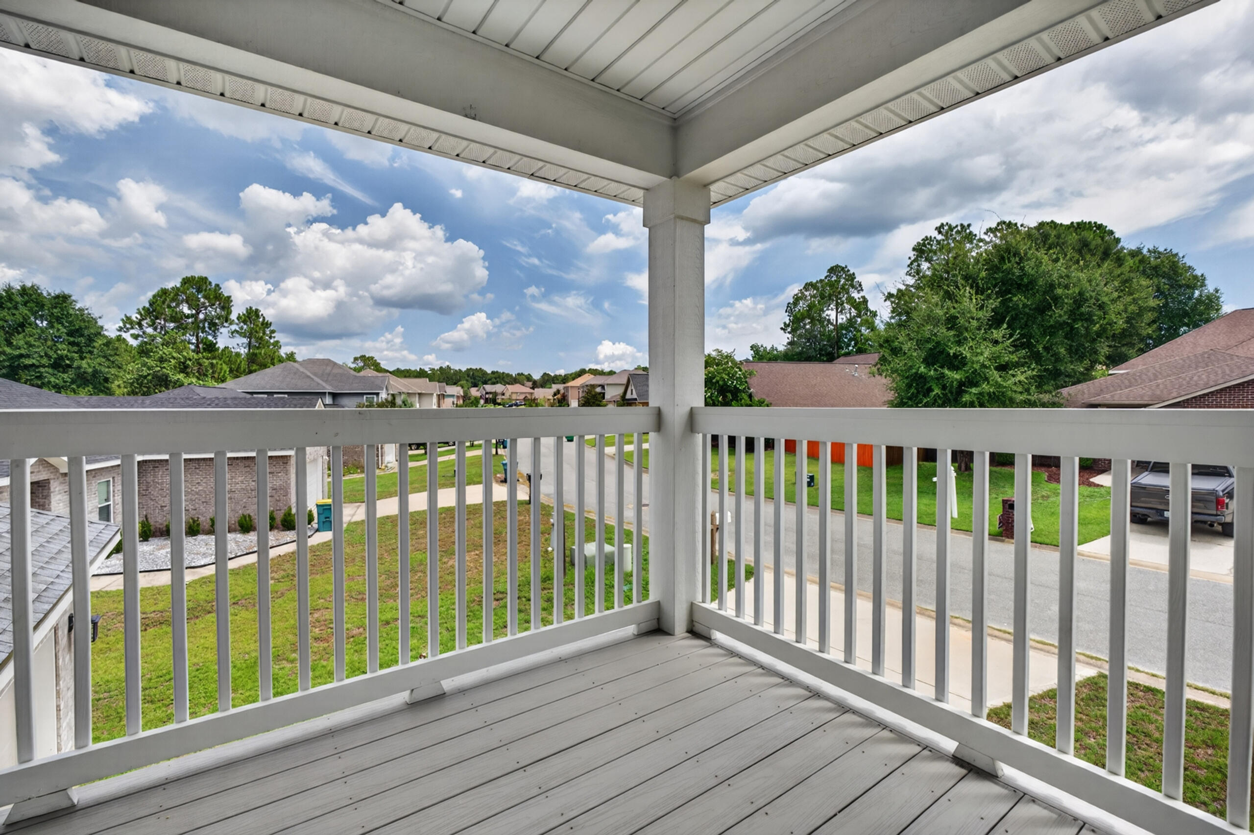 Covered balcony with gray decking and white railings overlooking a leafy residential street.