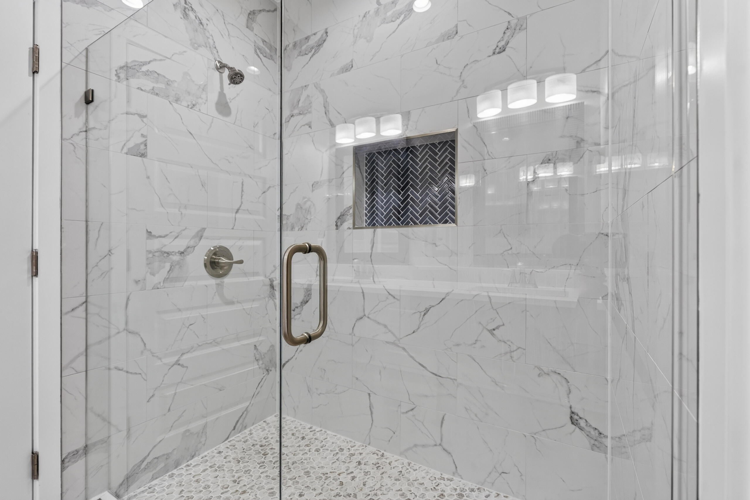 Walk-in shower with marble-look tile, dark herringbone niche, glass enclosure, and pebble mosaic floor.