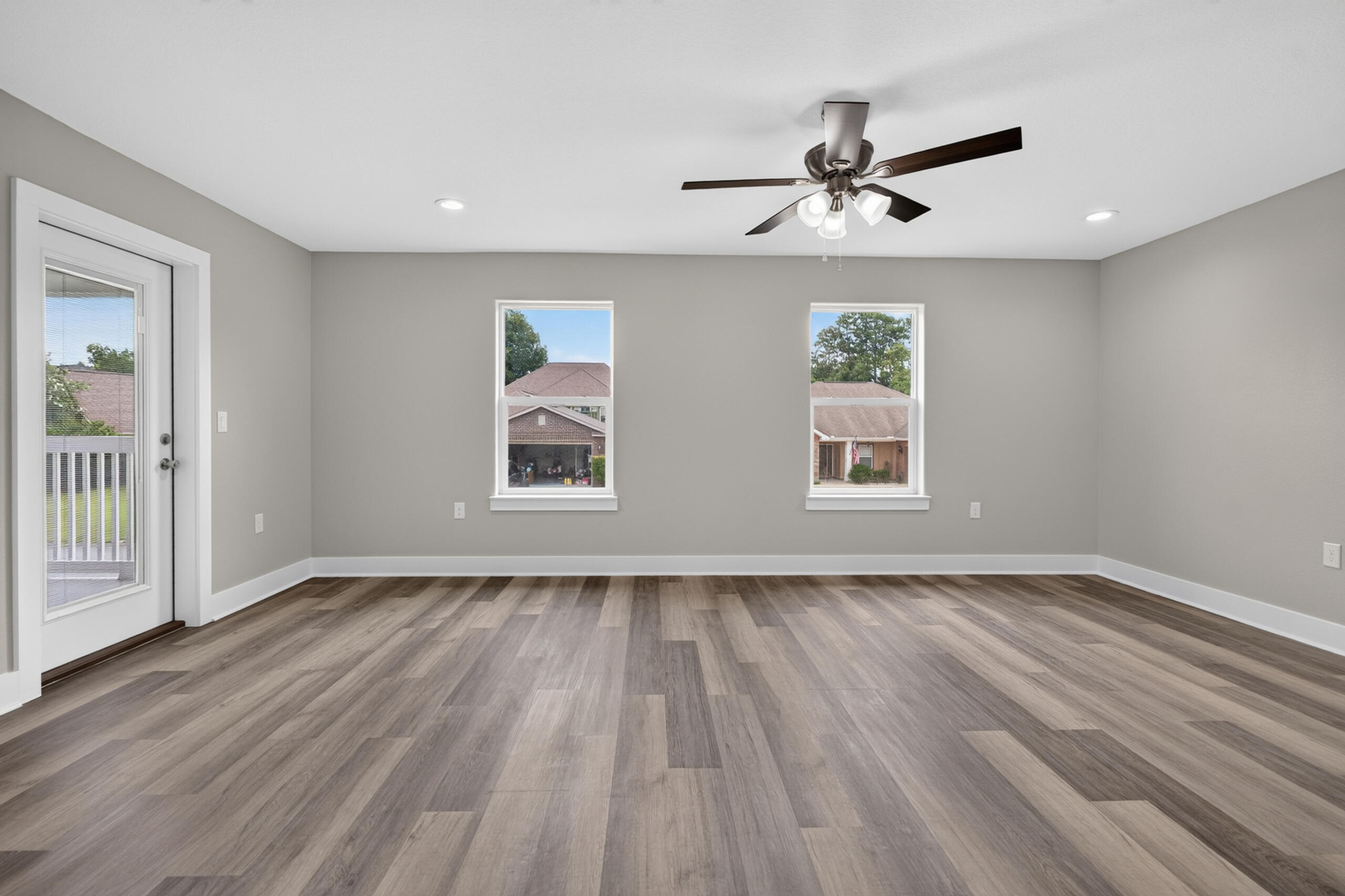 Empty living room with gray-brown plank flooring, a ceiling fan, two windows, and a glass-panel door.
