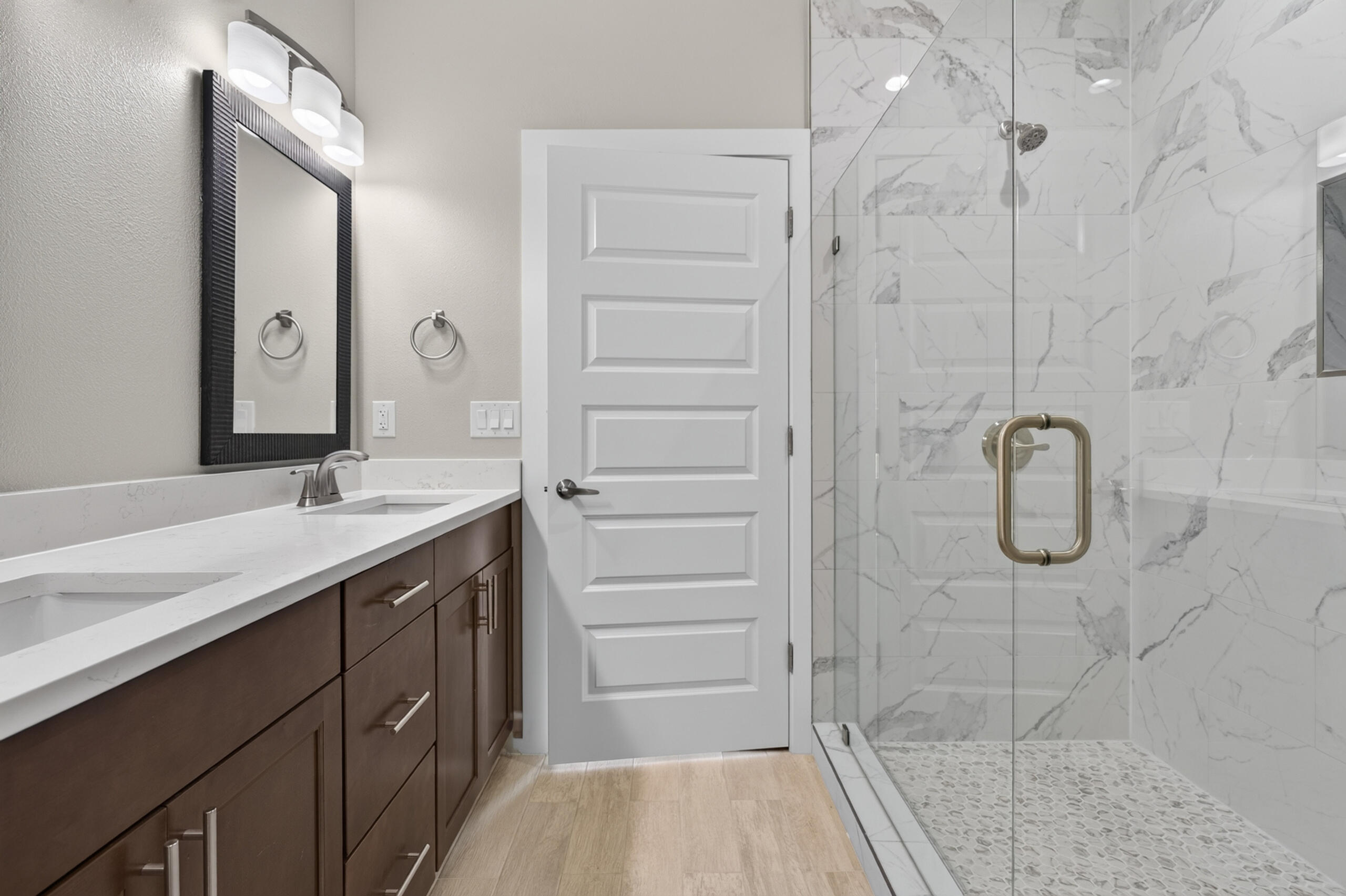 Bathroom with a dark wood vanity and glass shower featuring marble-look tile and pebble flooring.