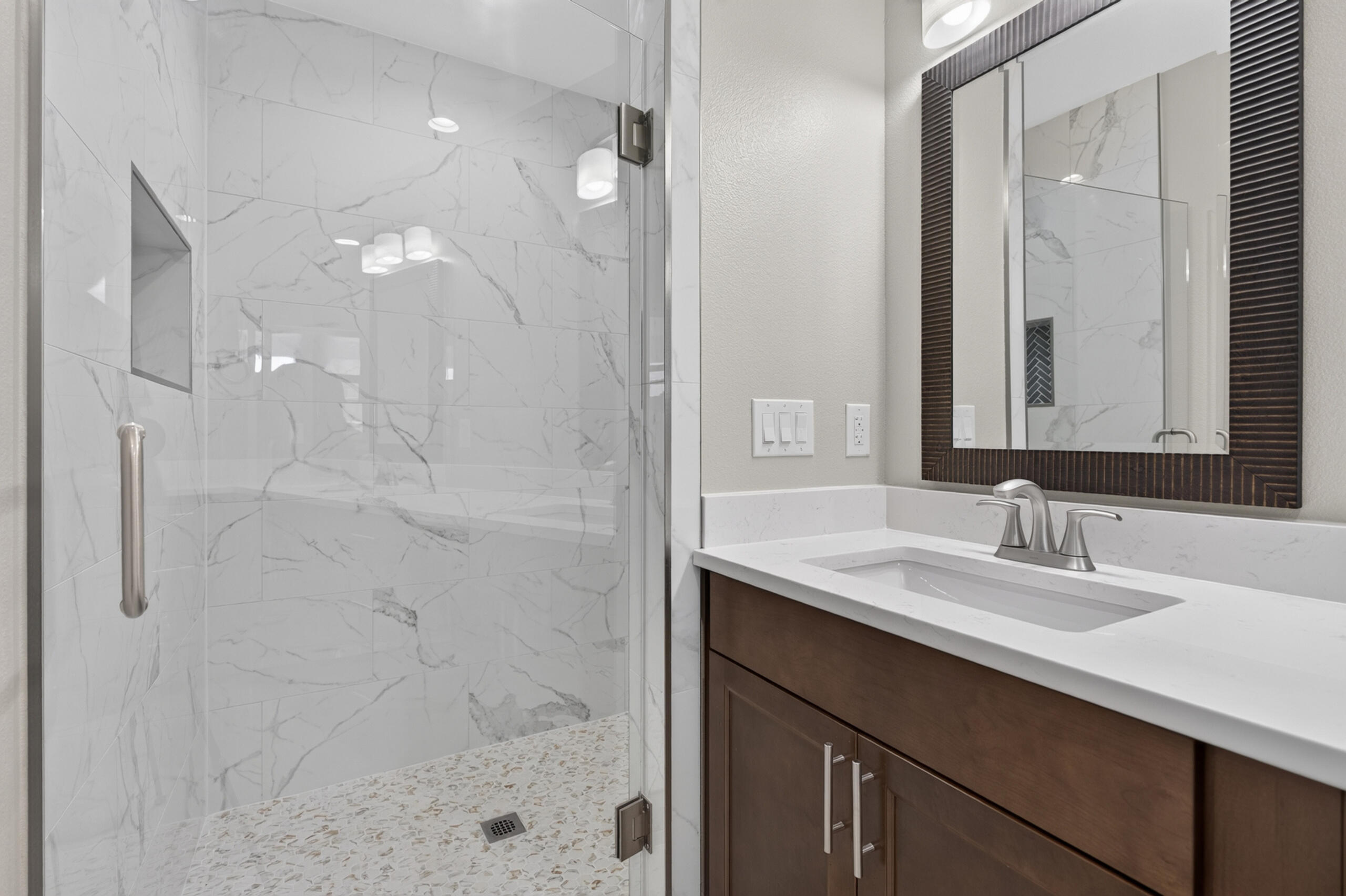 Bathroom with a marble-look tiled walk-in shower, pebble floor, and dark wood vanity.