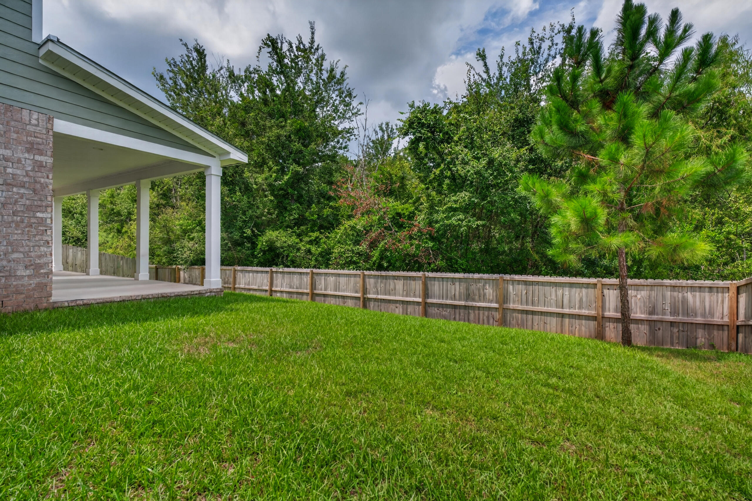Fenced grassy backyard with a covered patio and mature trees.