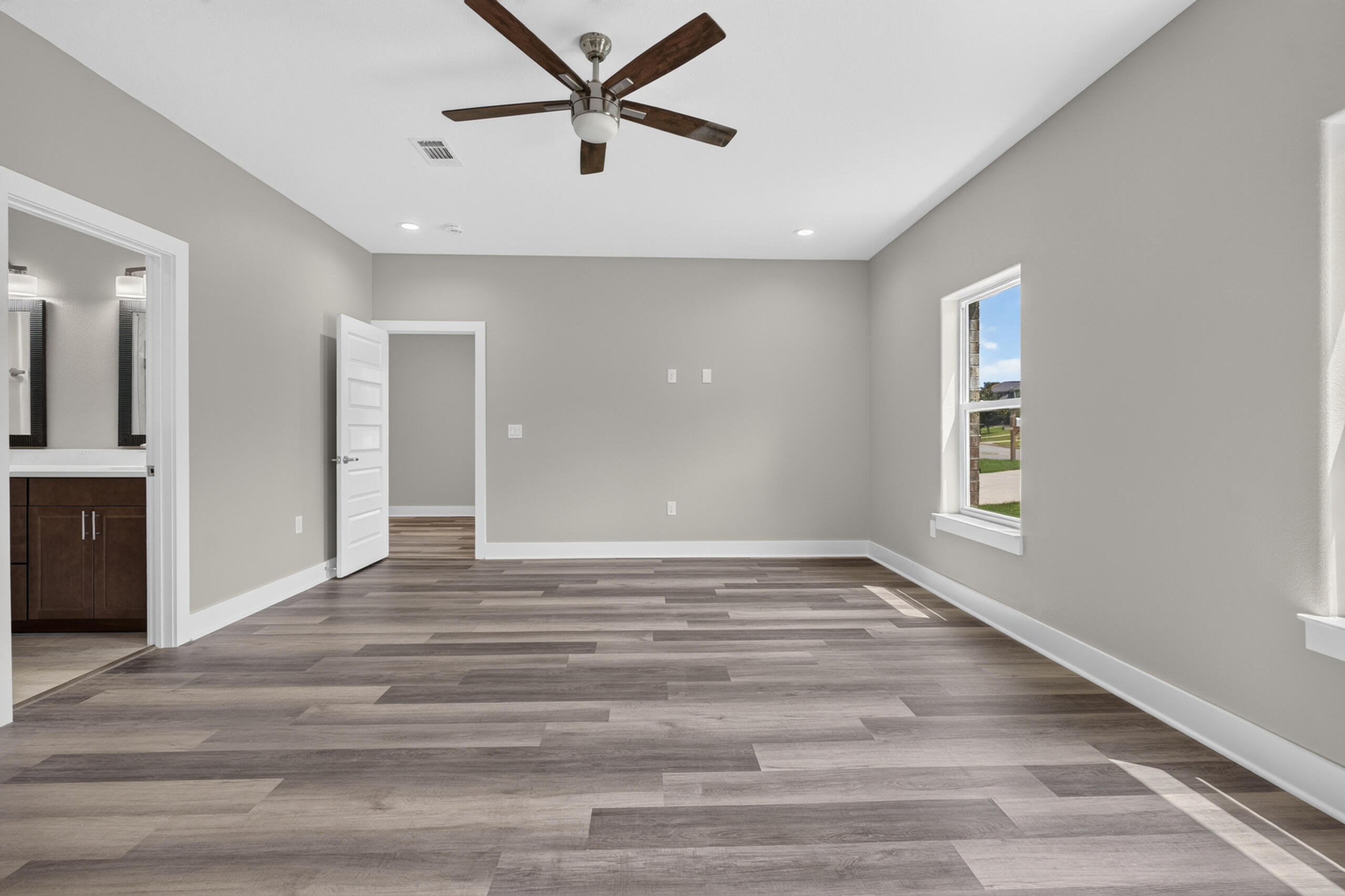 Empty bedroom with plank flooring, a ceiling fan, and an open doorway to a bathroom.