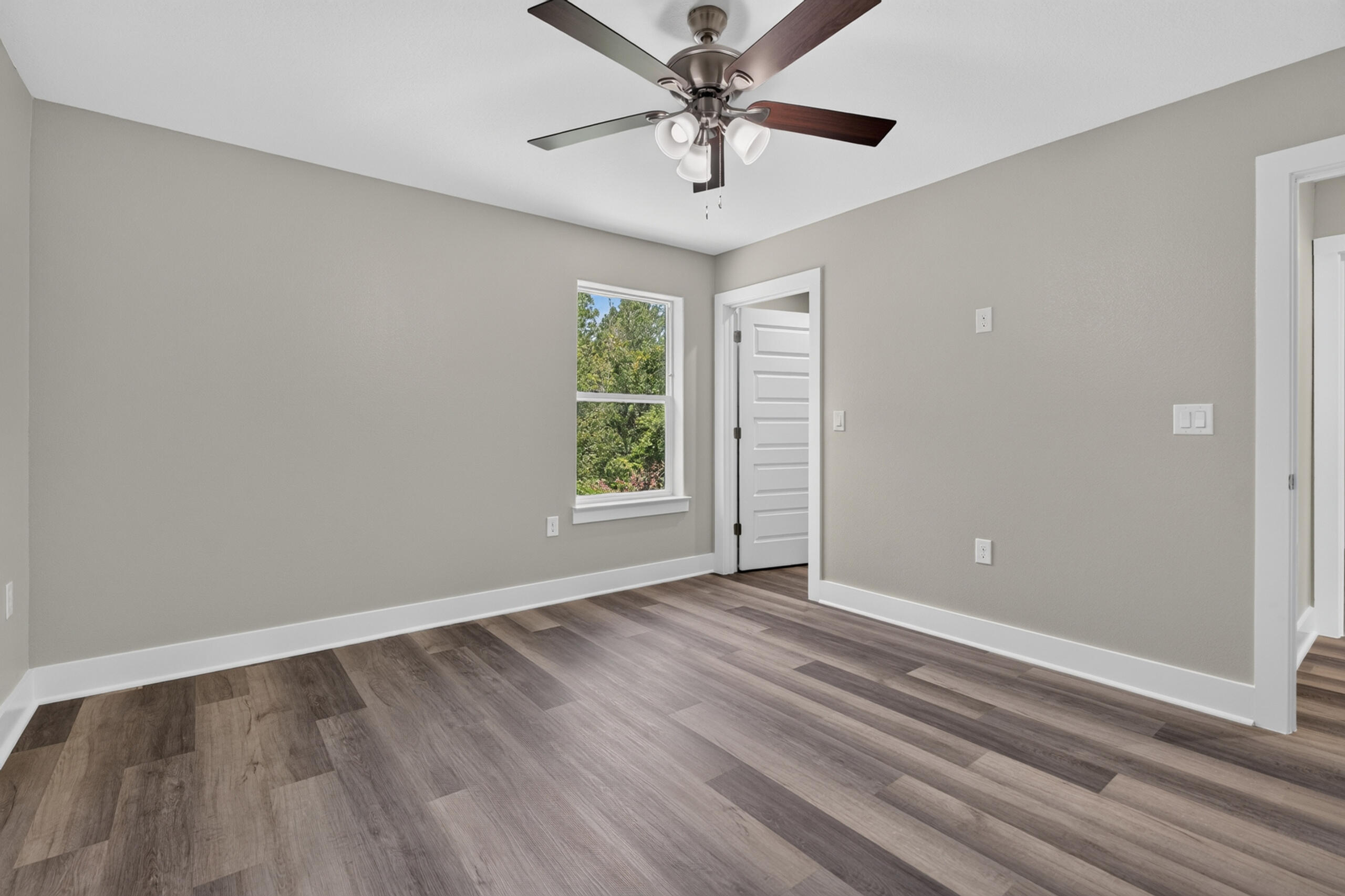 Empty bedroom with wood-look flooring, a ceiling fan, and a window overlooking trees.