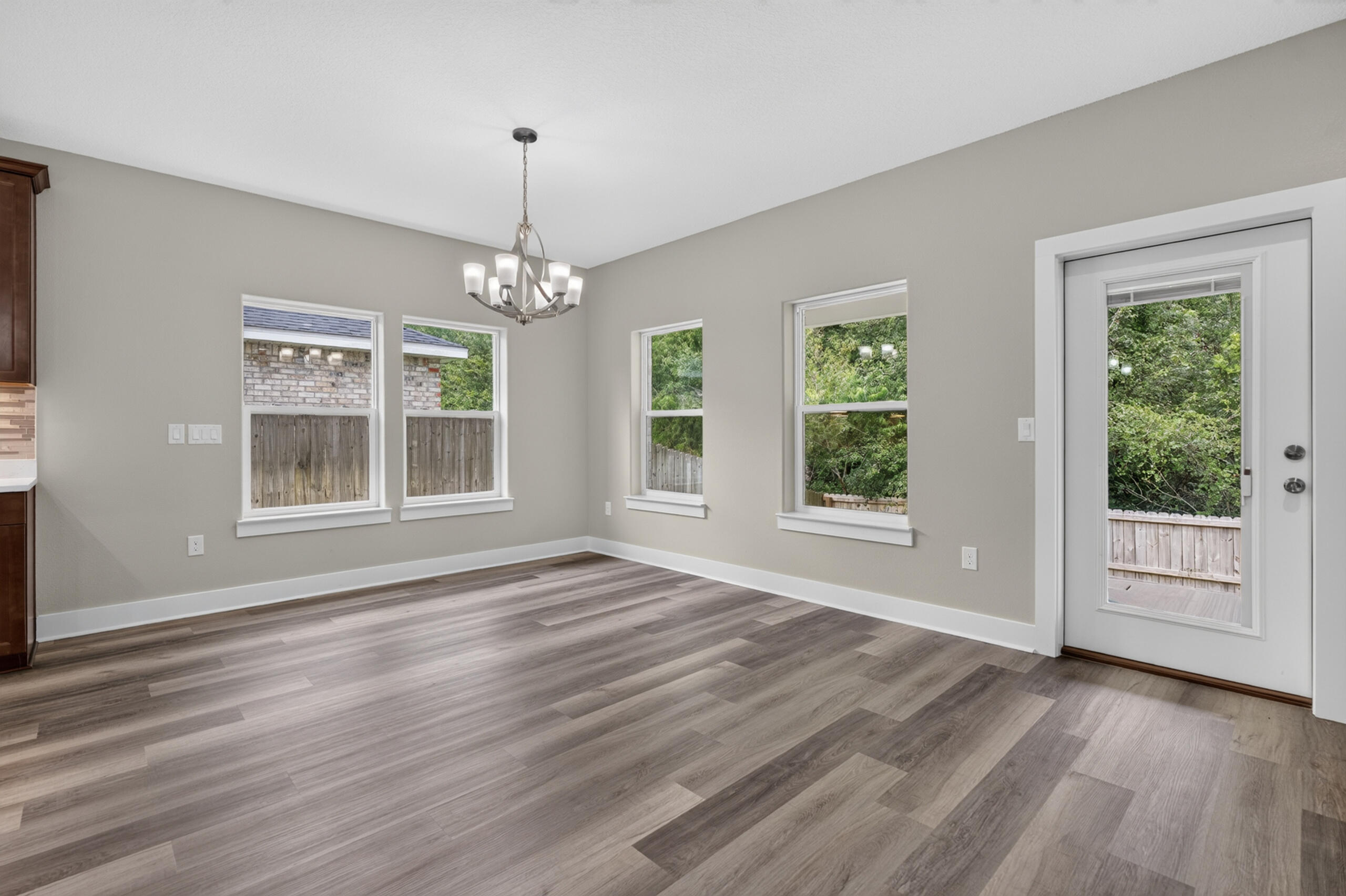 Empty dining area with gray wood-look floors, a chandelier, and windows beside a glass-paneled door.