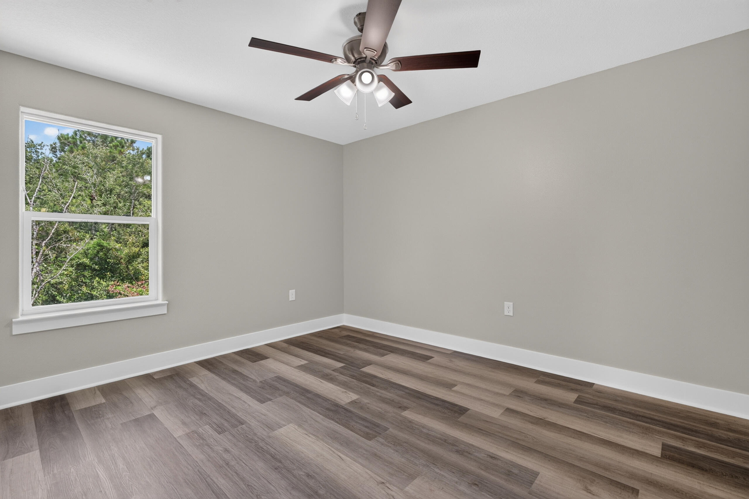 Empty bedroom with a ceiling fan, wood-look flooring, and a window overlooking trees.