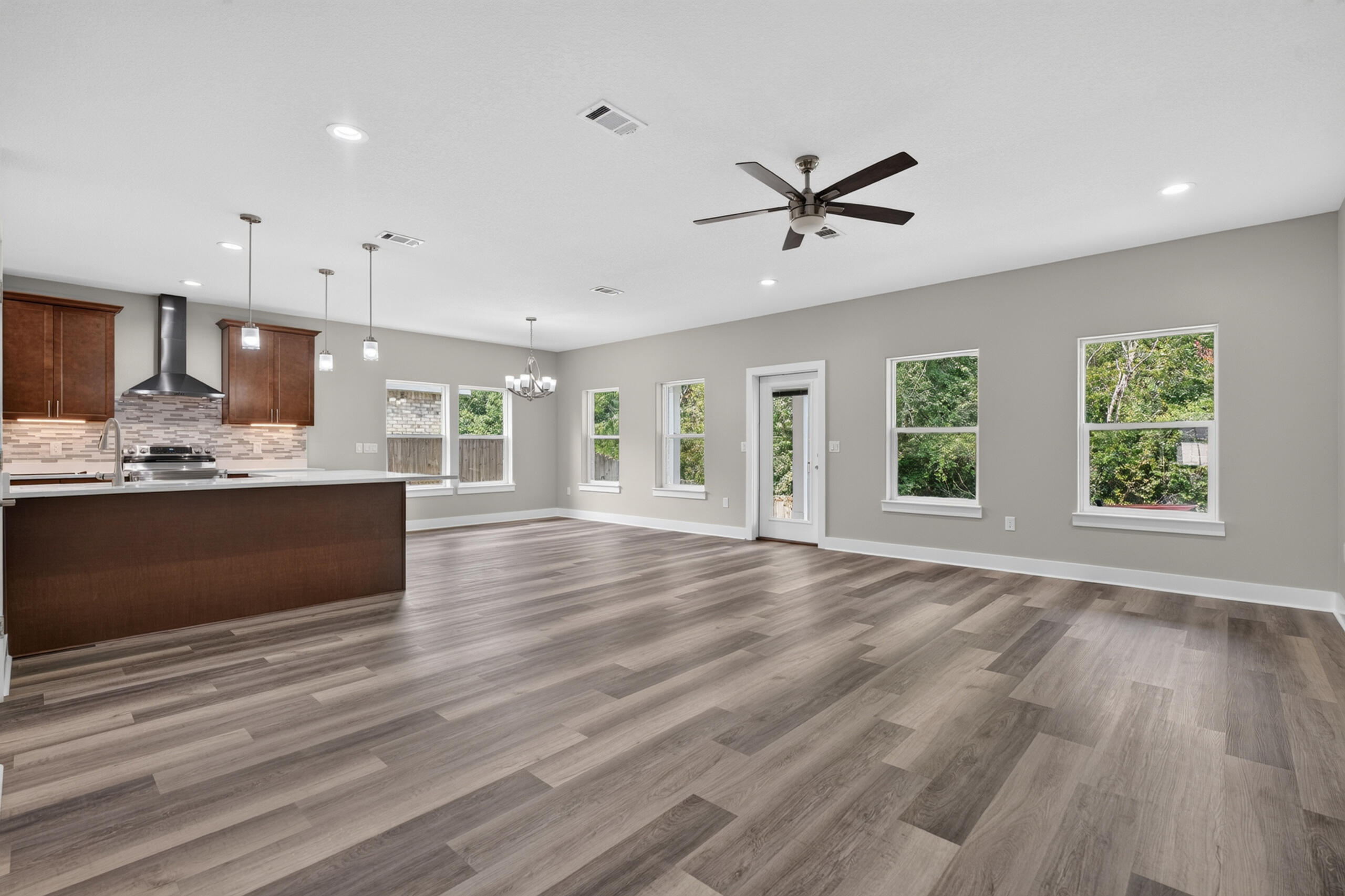 Open-plan kitchen and empty dining area with dark wood island, plank flooring, and windows overlooking greenery.