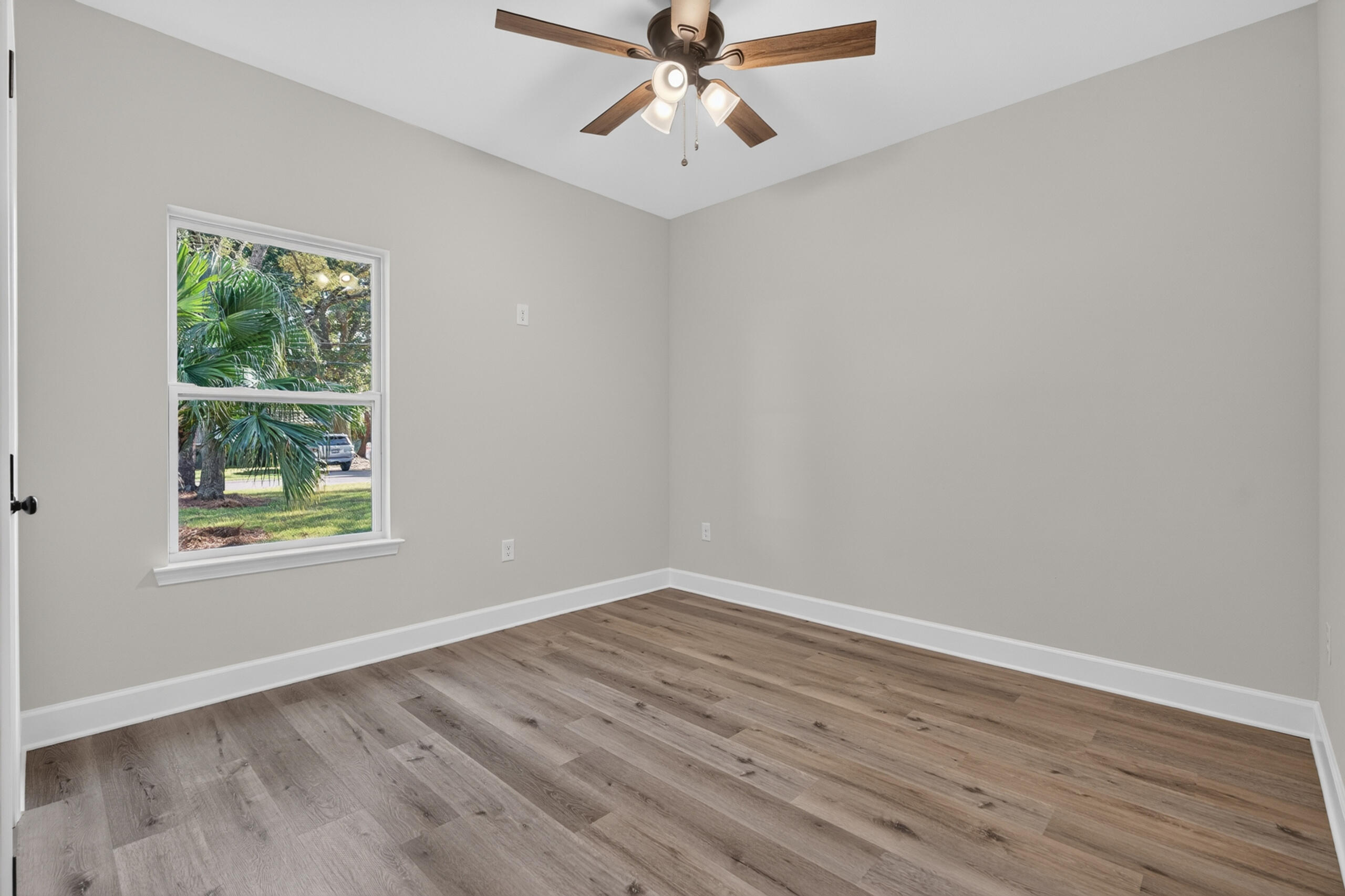 Empty room with wood-look flooring, a ceiling fan, and a window overlooking leafy landscaping.