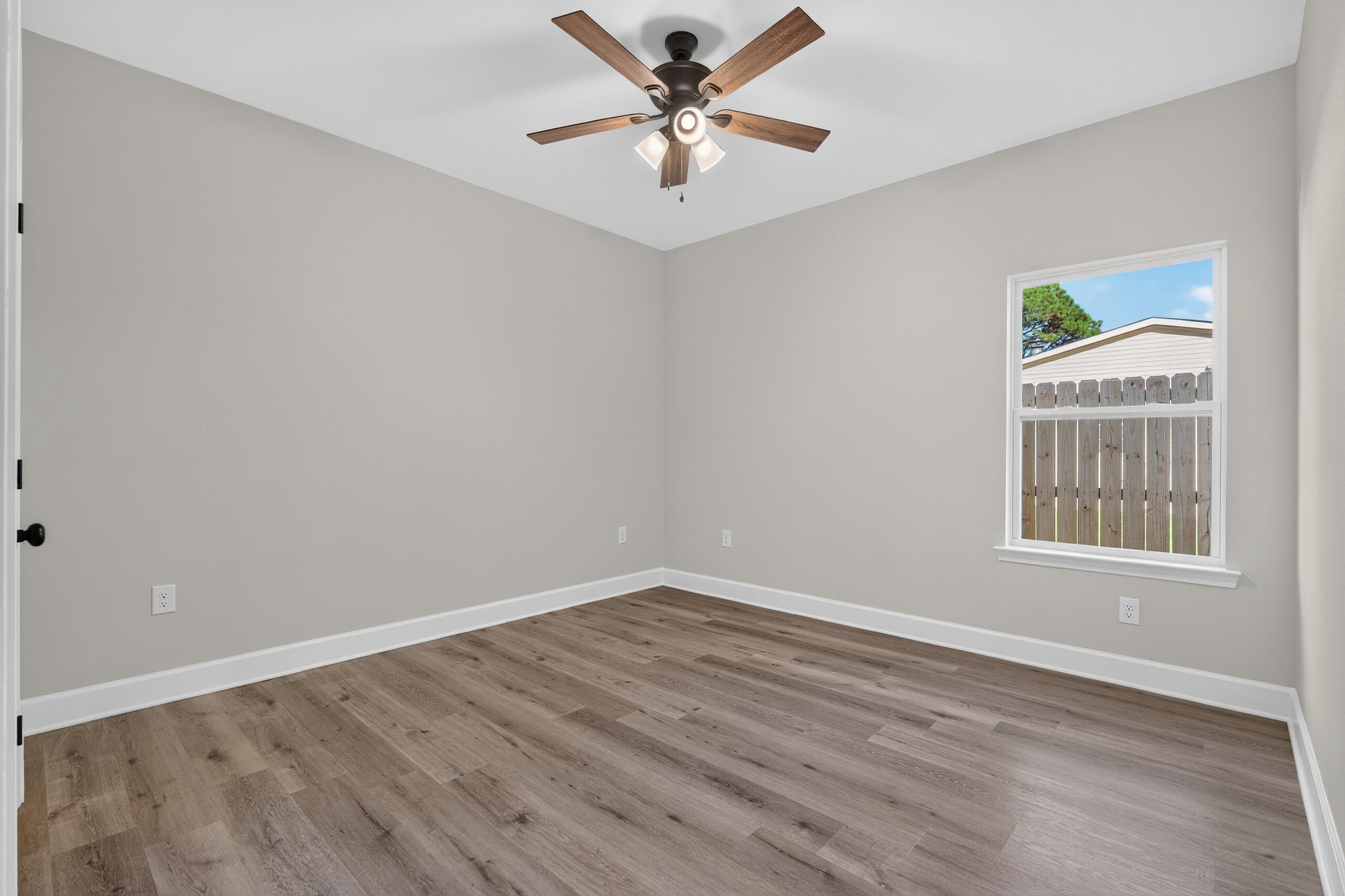 Empty bedroom with wood-look flooring, ceiling fan, and a window facing a wooden fence.