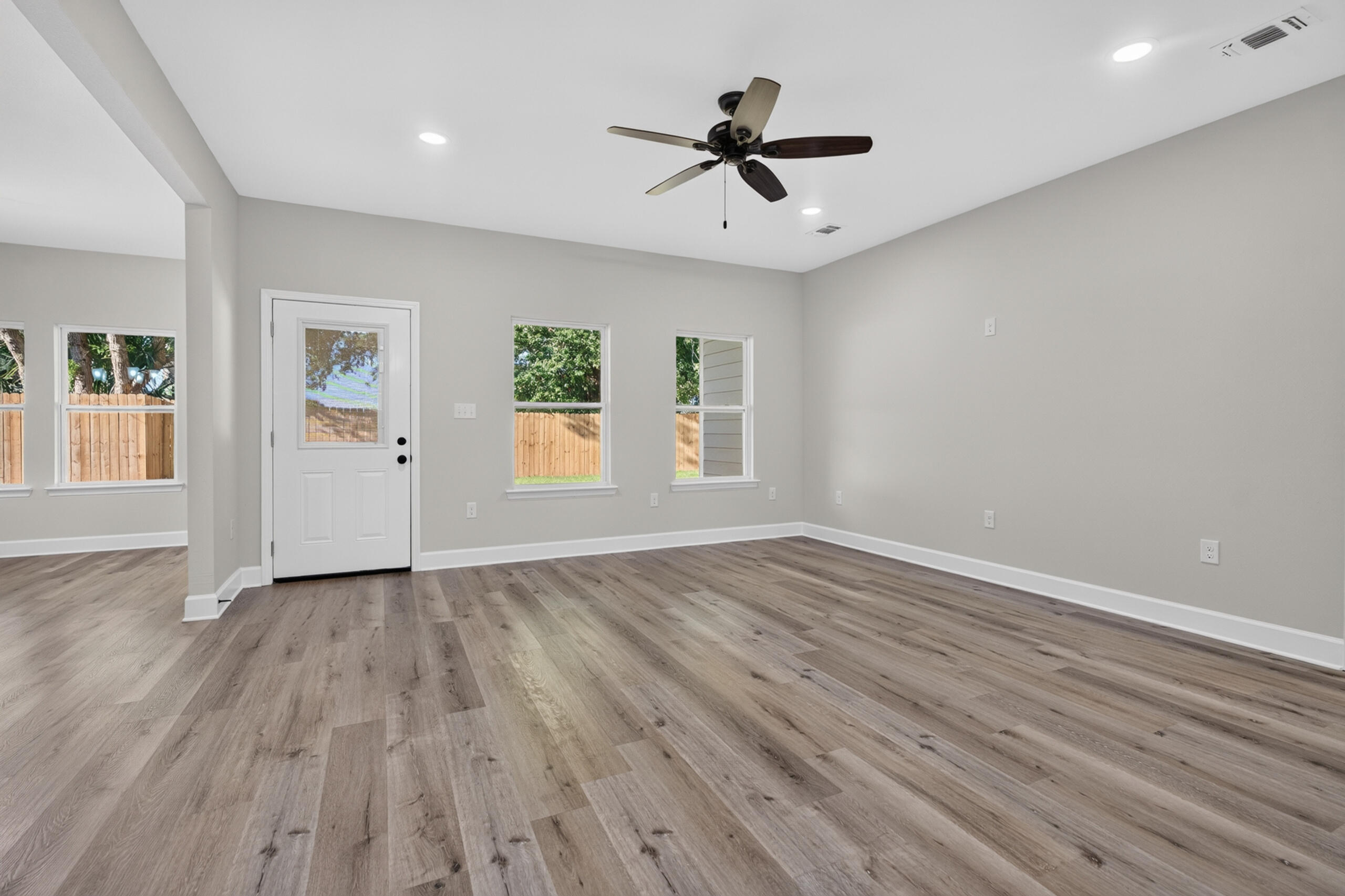 Empty room with wood-look flooring, a ceiling fan, and windows facing a fenced yard.