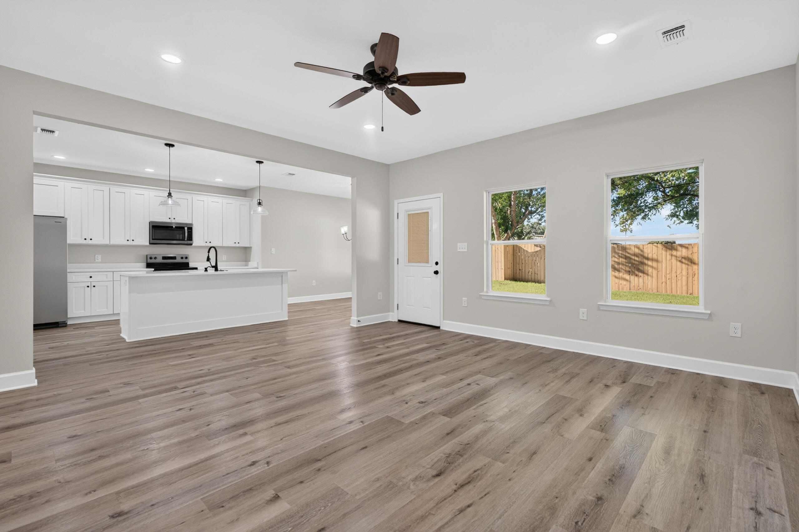 Open living area with plank flooring, a white kitchen island, and windows facing a fenced yard.