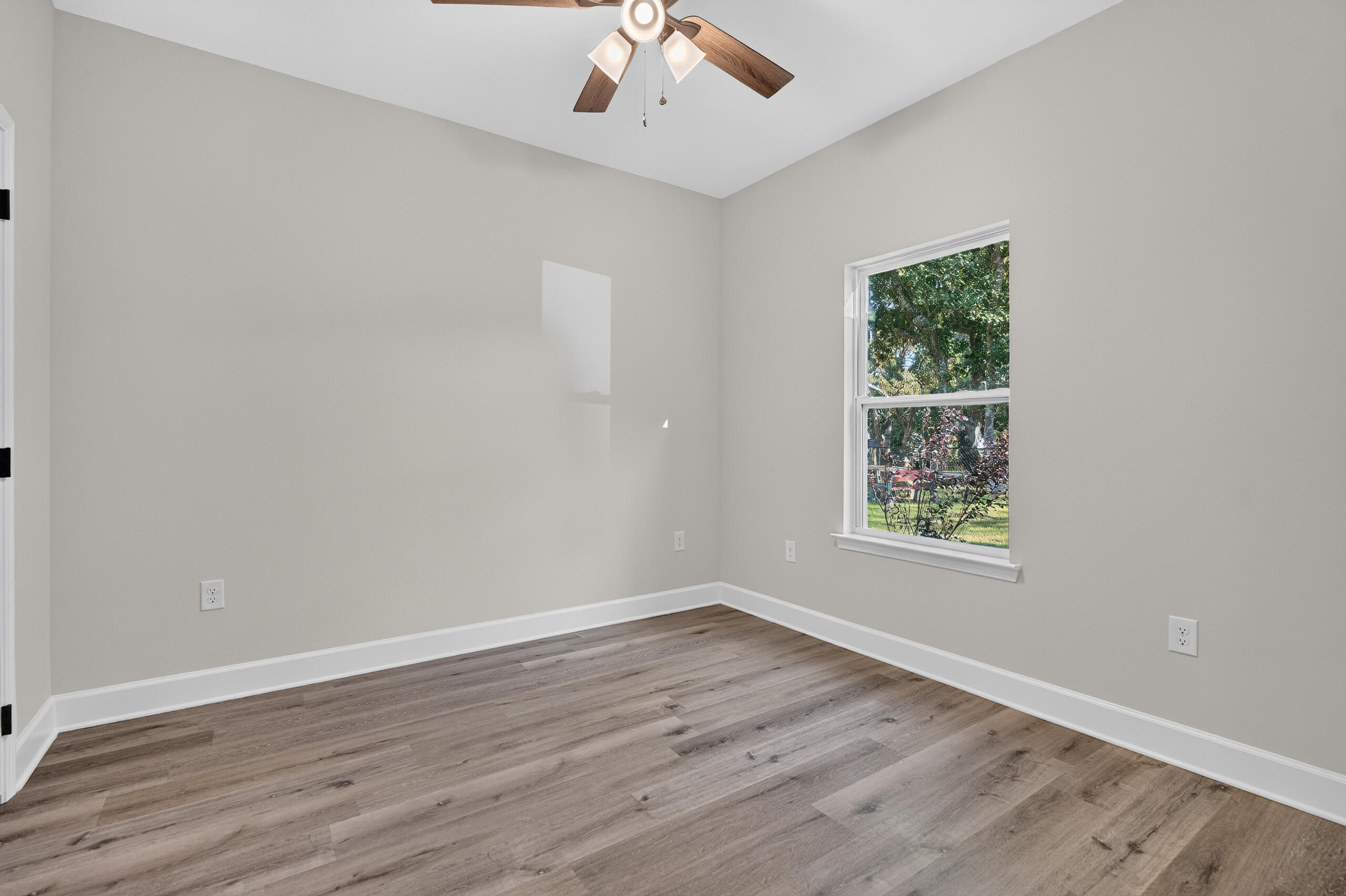 Empty bedroom with wood-look flooring, a ceiling fan, and a window overlooking trees.