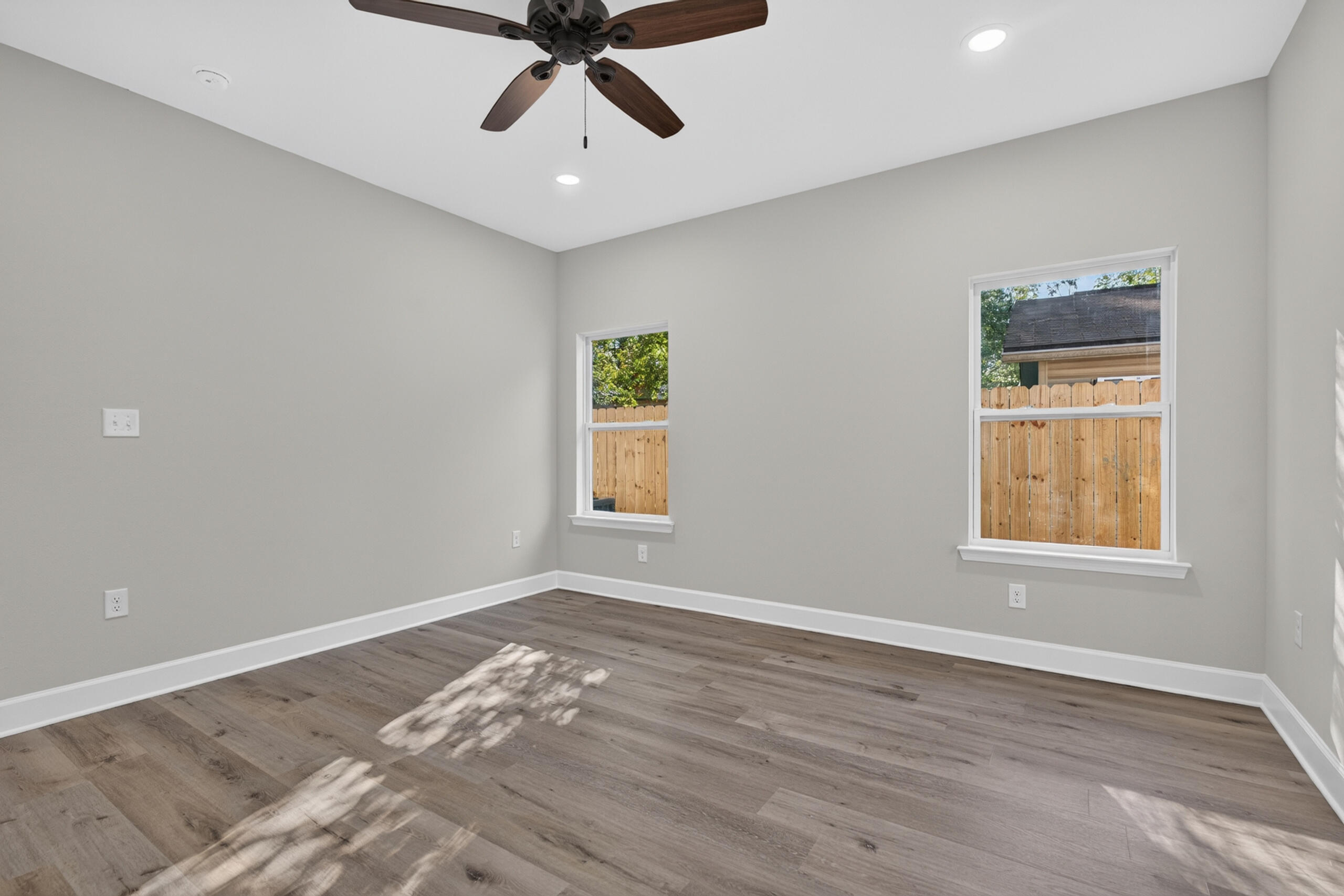 Empty bedroom with wood-look floors, a ceiling fan, and two windows facing a wooden fence.